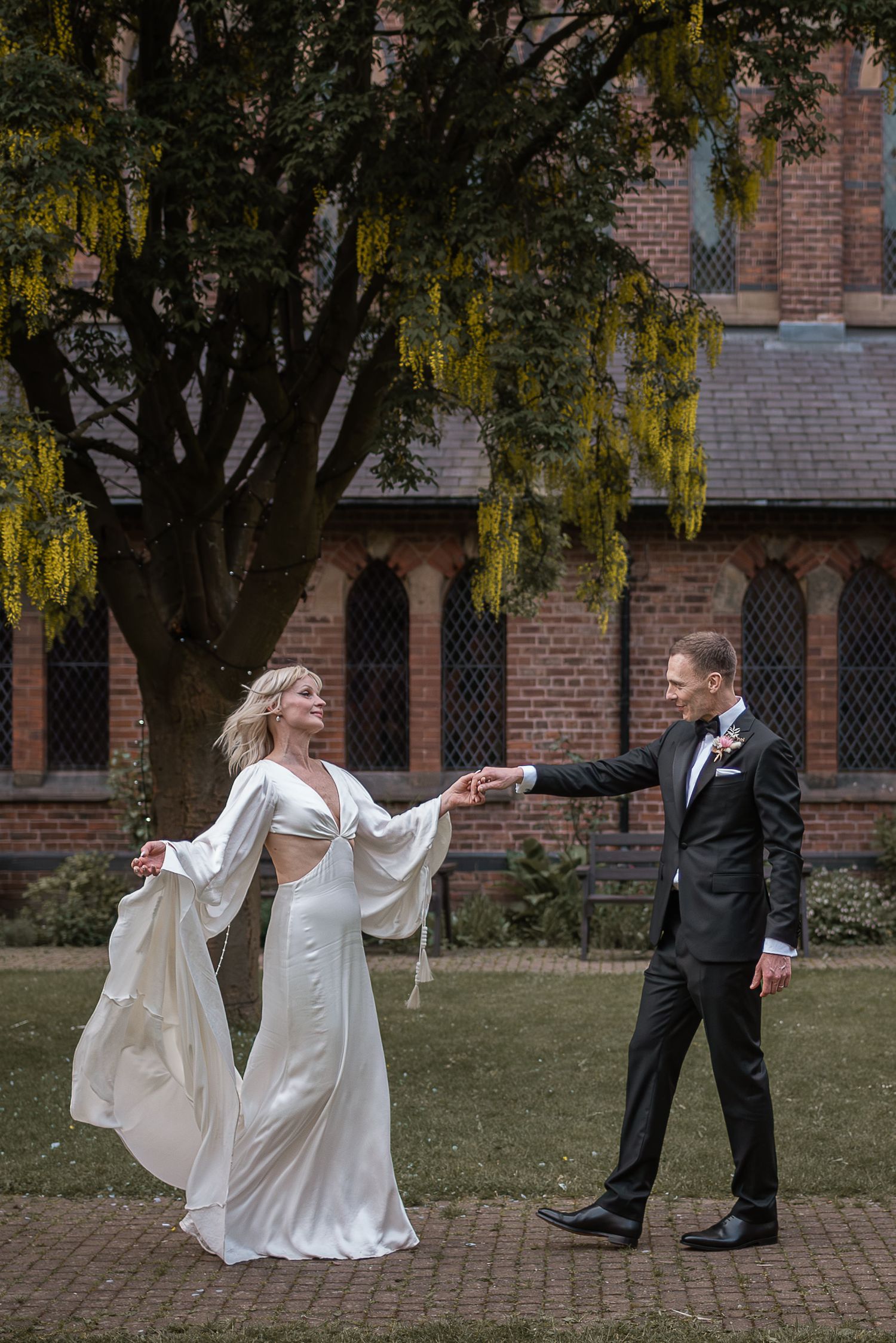 A couple dances playfully on a brick path in front of a historic church with hanging yellow flowers.