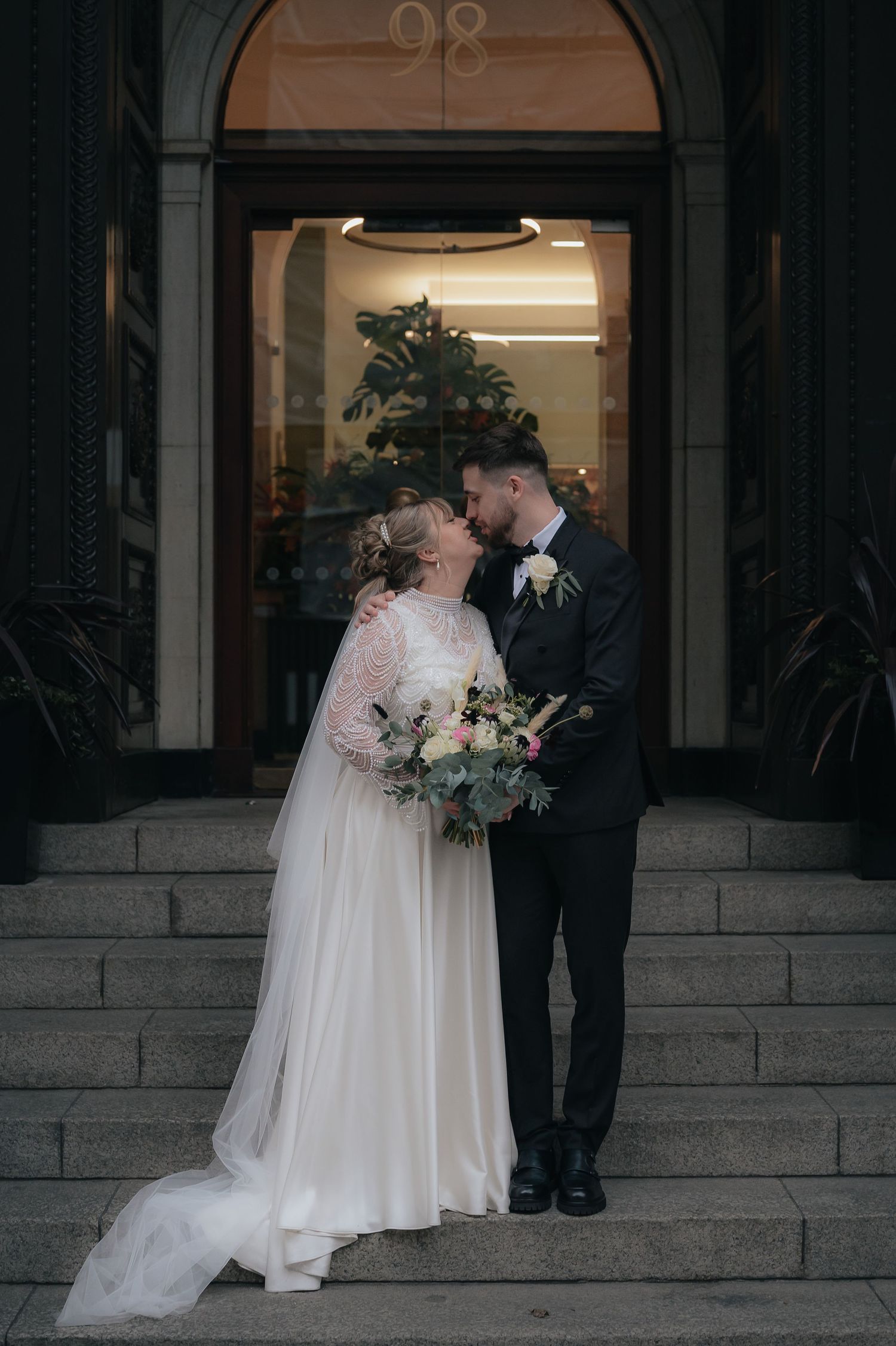 A couple shares an intimate moment on stone steps in front of an arched doorway at their wedding celebration.