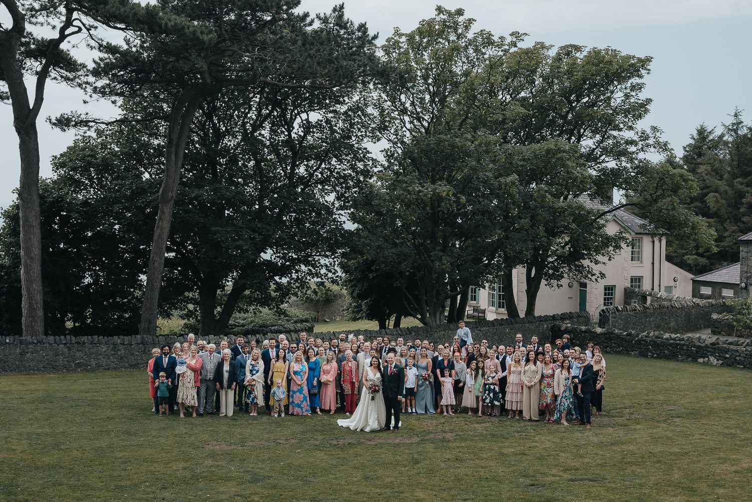 Large wedding party gathered on a grassy lawn with trees and stone walls in the background for a group photo.
