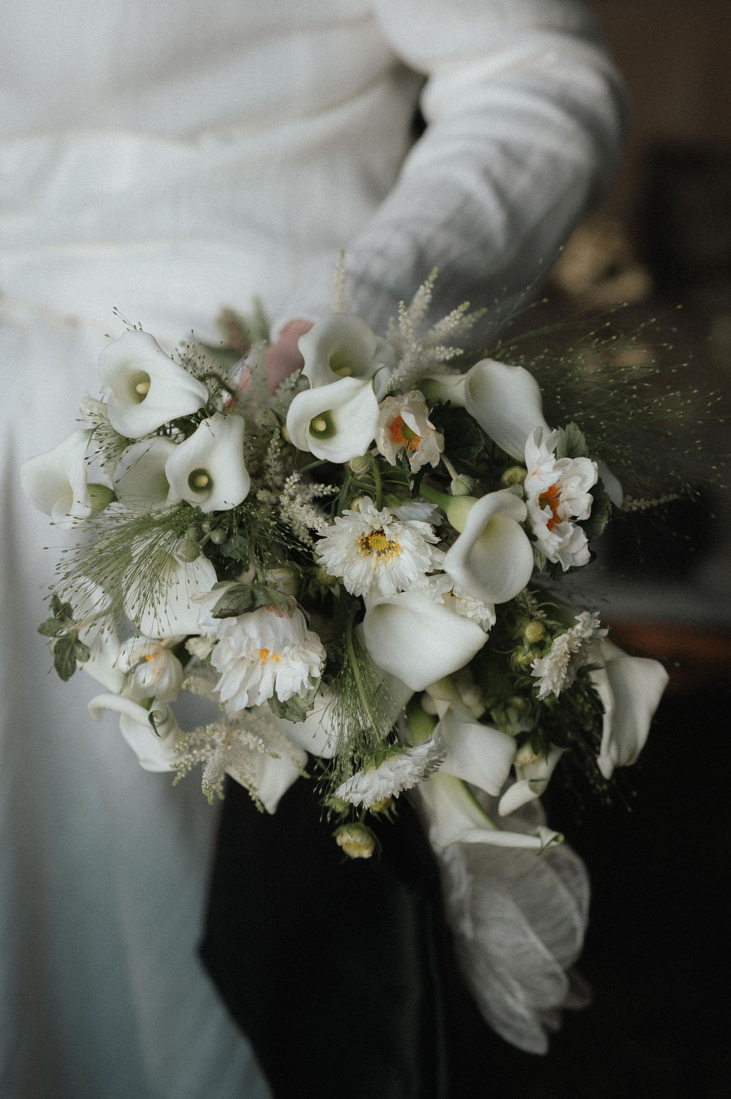 Delicate white floral arrangement with calla lilies and textured greenery against a dark background.