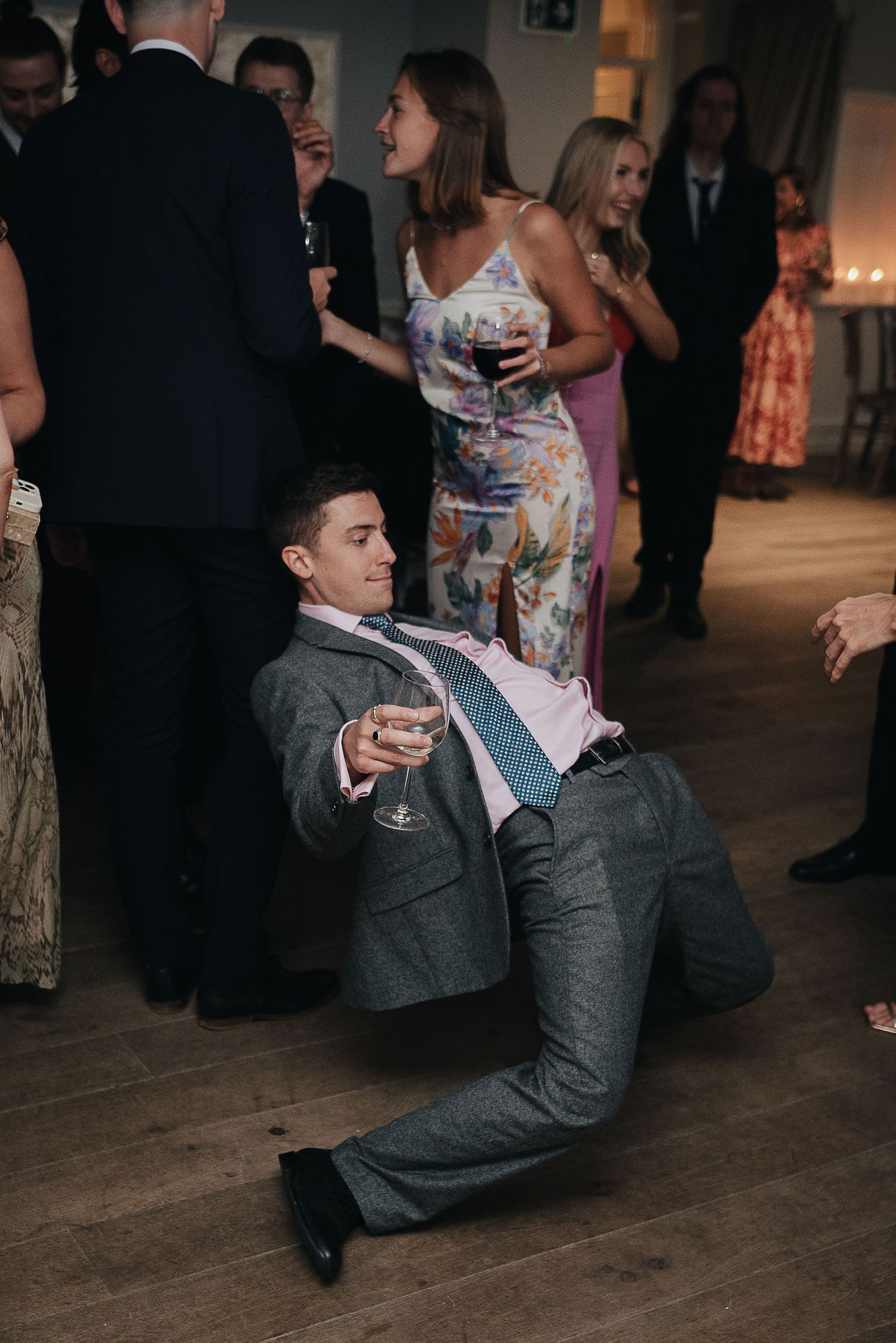 Wedding guest in grey suit performs dramatic dance move while holding a drink at reception party.
