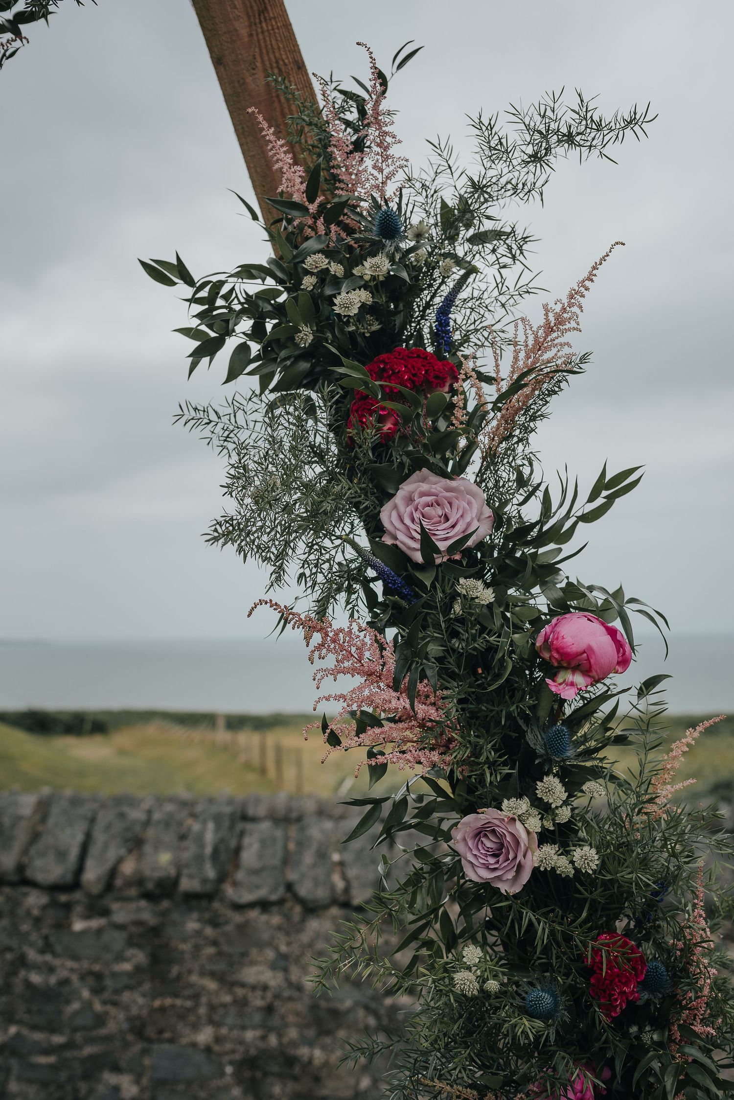 Romantic floral arrangement with pink roses and greenery climbing up a wooden post against cloudy sky.