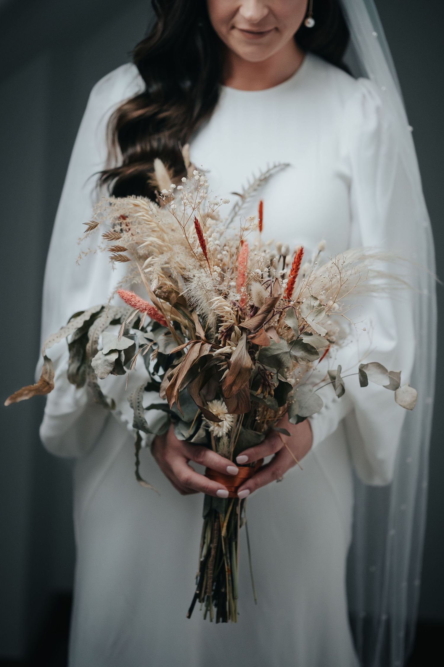 Elegant bridal bouquet with dried flowers and pampas grass held against a white wedding dress.