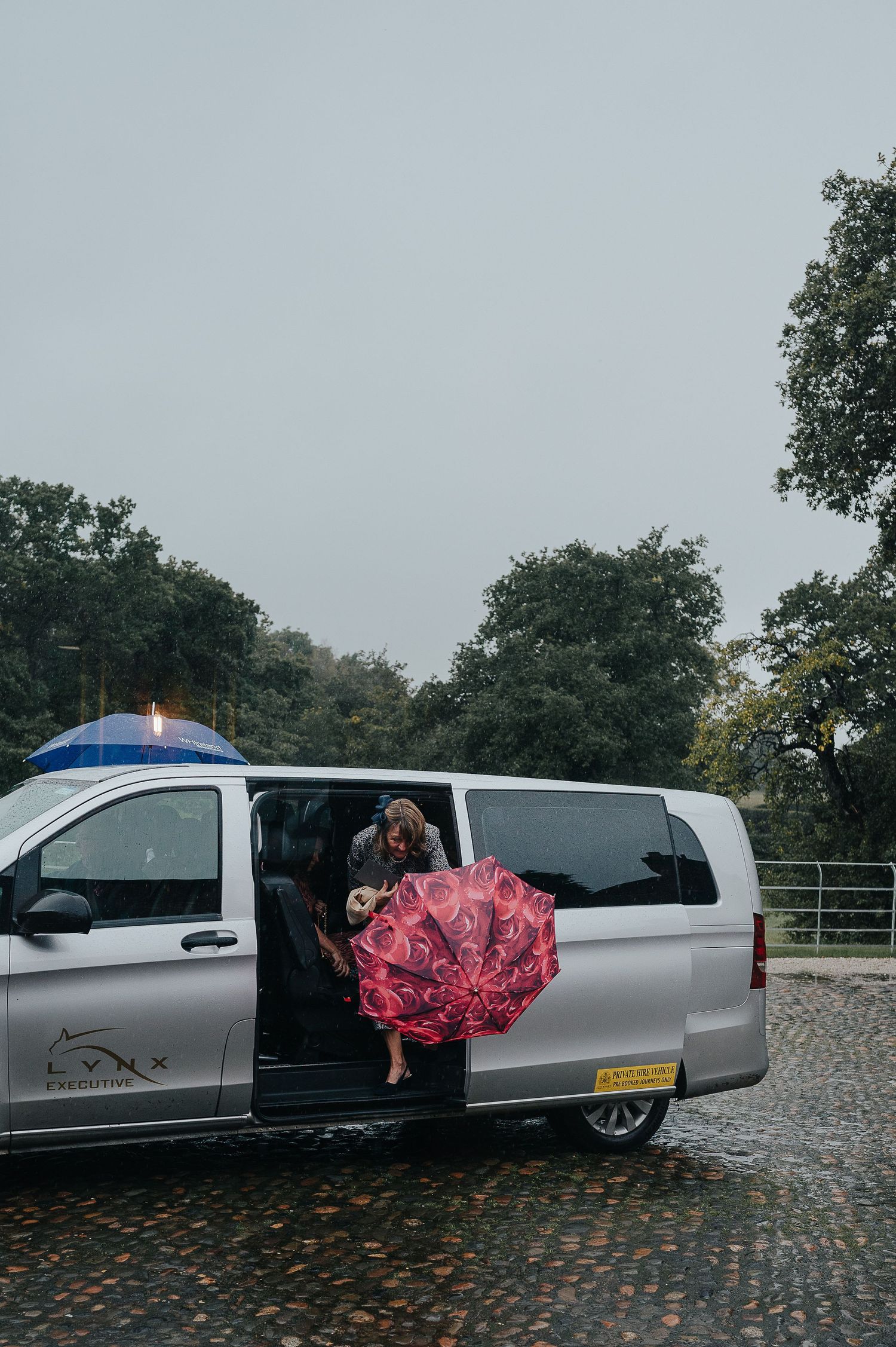 Someone exiting a silver van with a red umbrella on a rainy day.