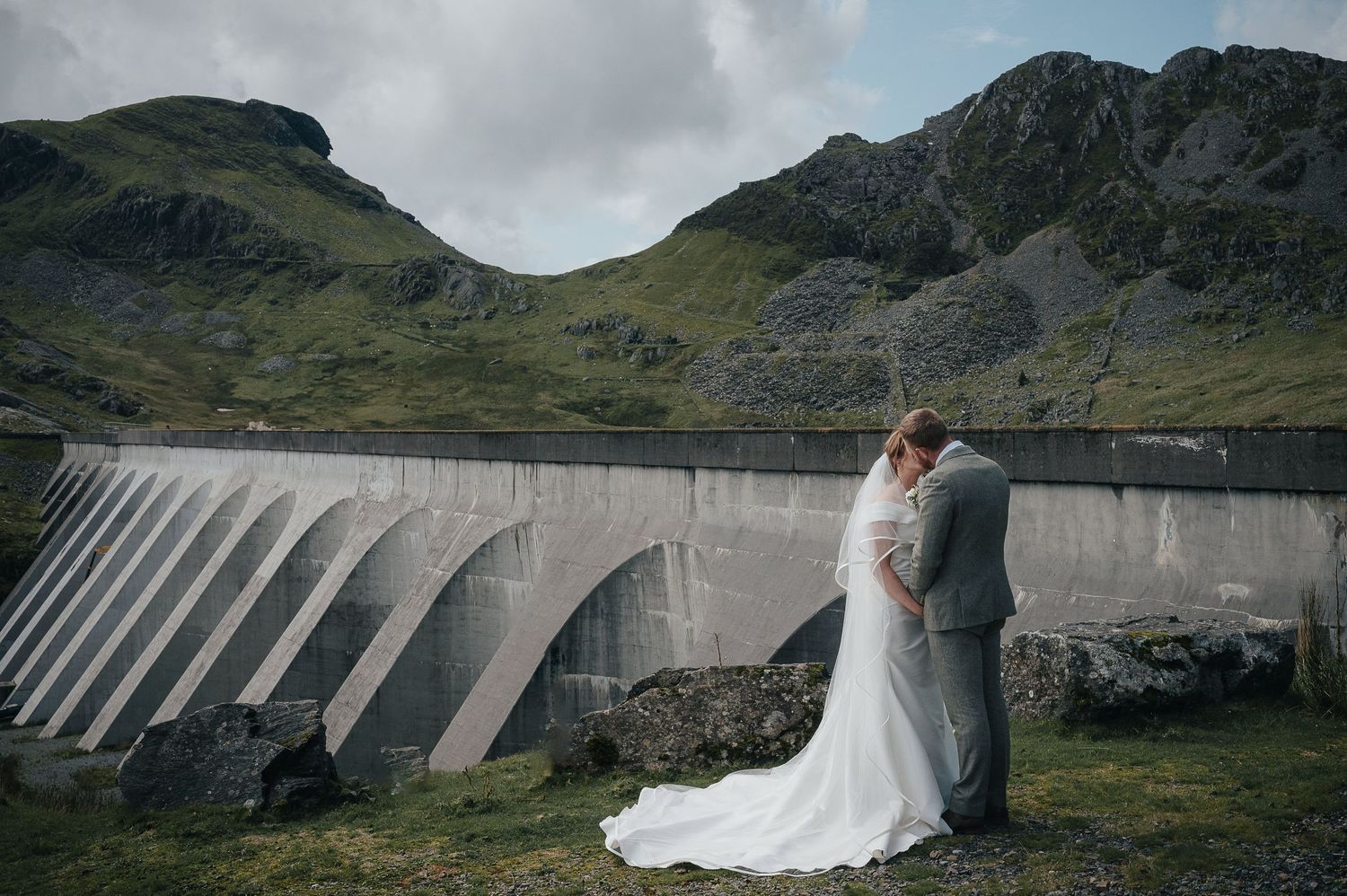A couple shares a romantic moment in front of a dramatic concrete dam in mountainous terrain.