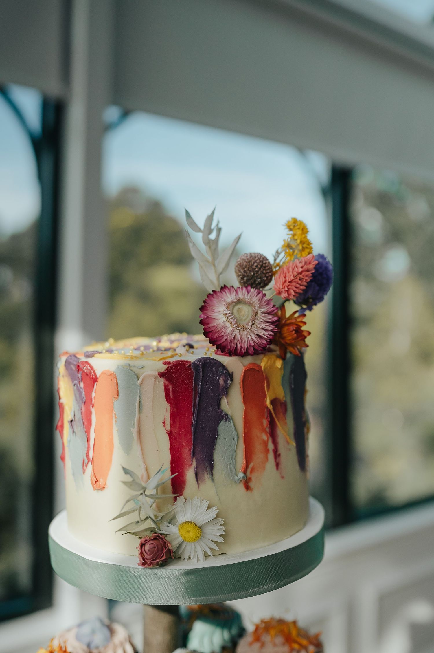 A modern wedding cake decorated with colorful drip design and fresh flowers on a mint green stand.