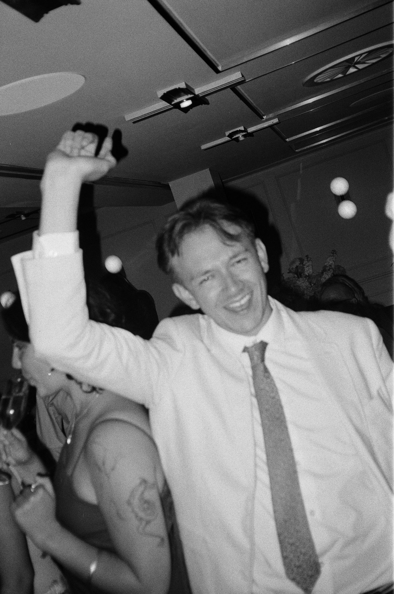 Man in white shirt and tie raising fist in celebration at a party, black and white photo.