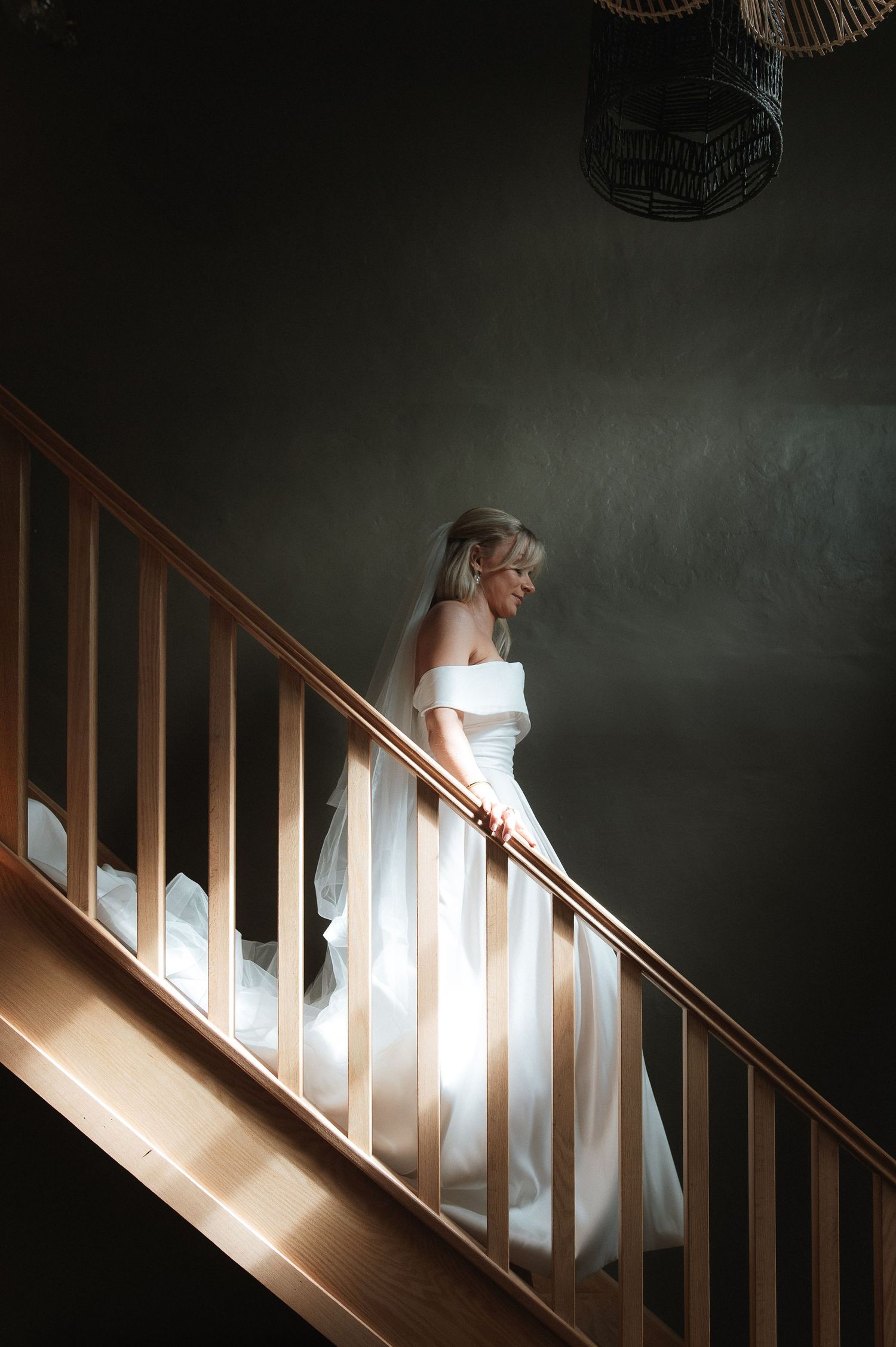 A figure in an elegant white wedding dress descends a dramatically lit wooden staircase.Blackton Grange Wedding. Luke Hassall Photography.