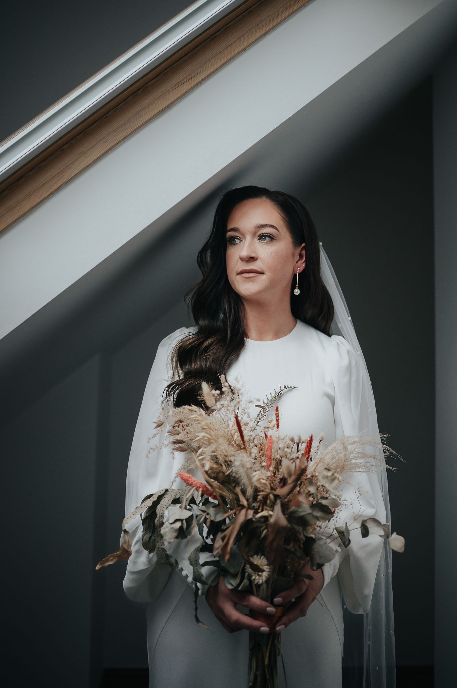 A bride in a long-sleeve white dress holds a bouquet of dried flowers and pampas grass against a gray wall.