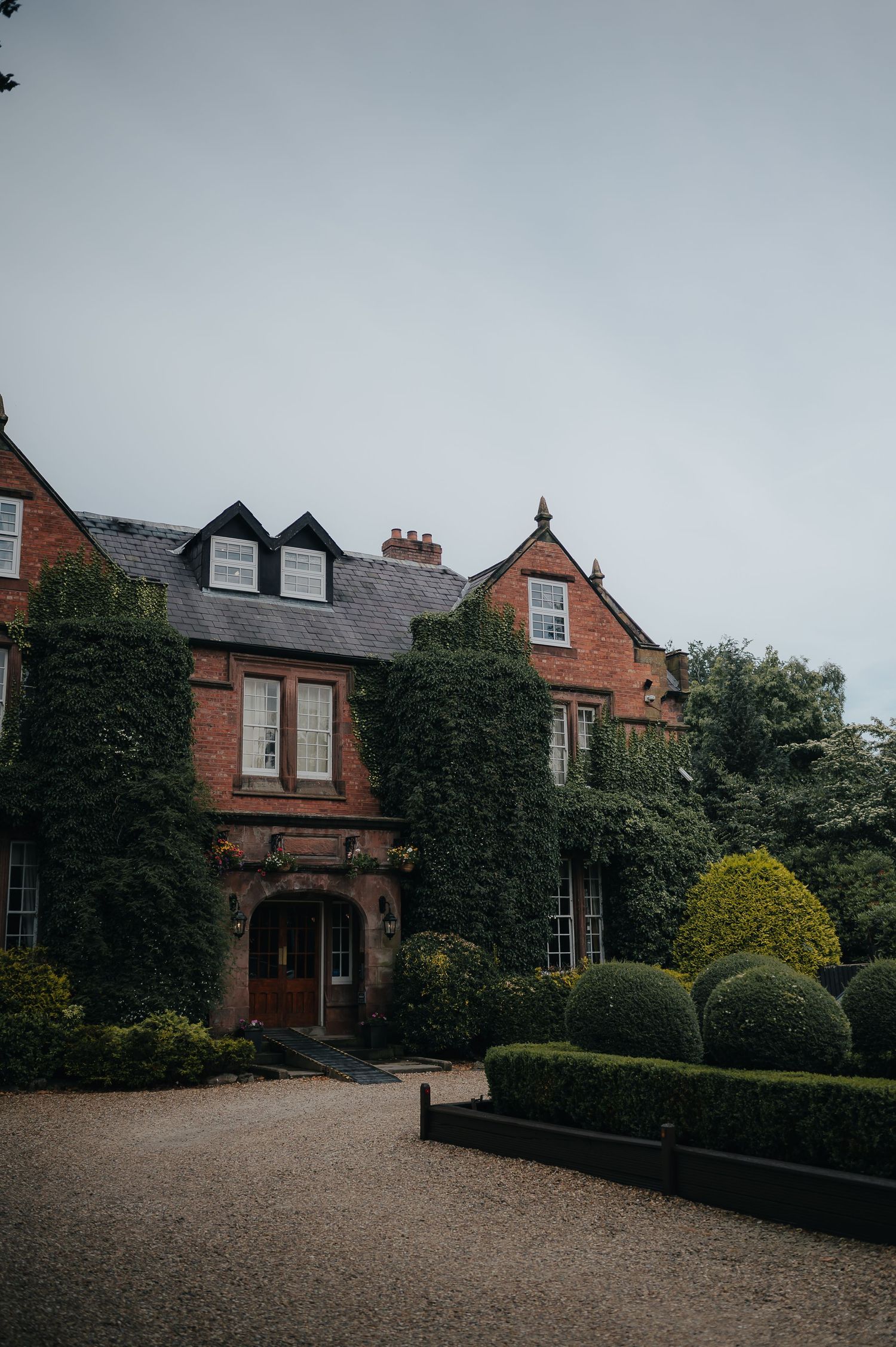 An ivy-covered brick manor house with dormers and manicured hedges on a cloudy day.