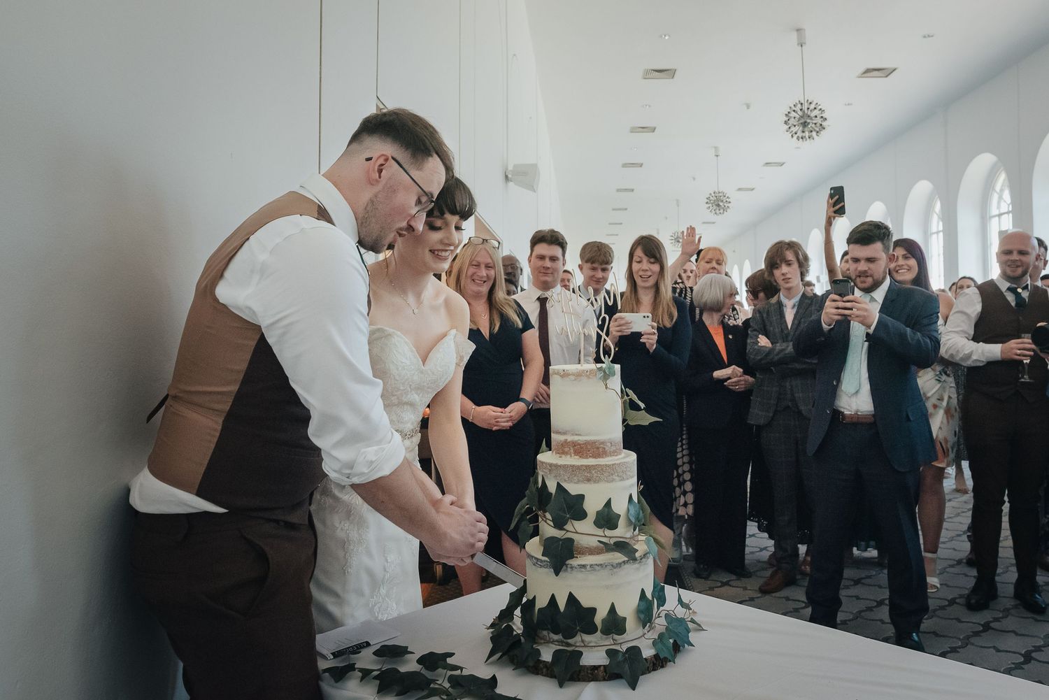 Wedding cake cutting moment surrounded by celebrating guests in a bright indoor venue.