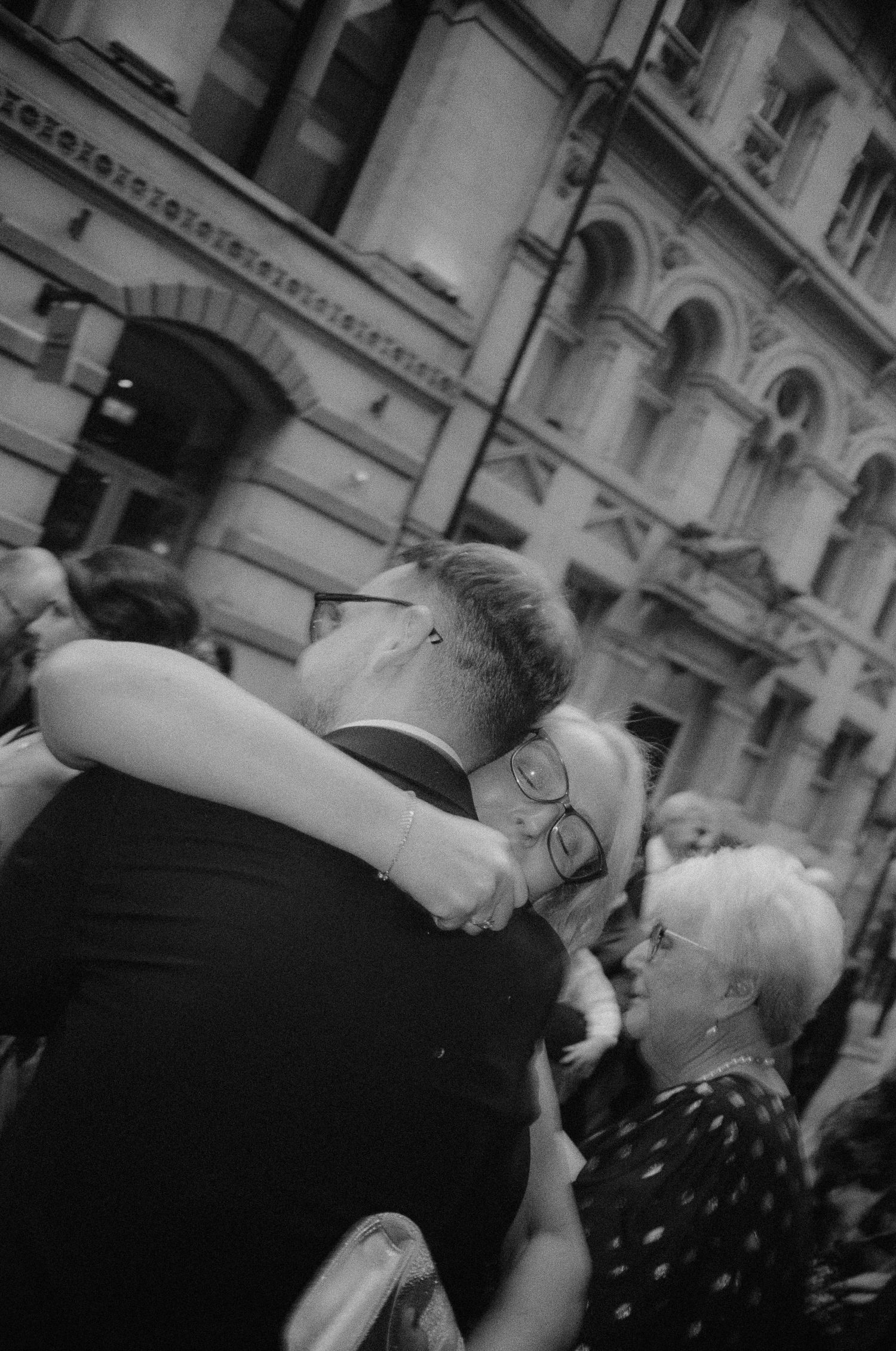 Black and white photo of two people embracing on a busy city street, with classic architecture in the background.