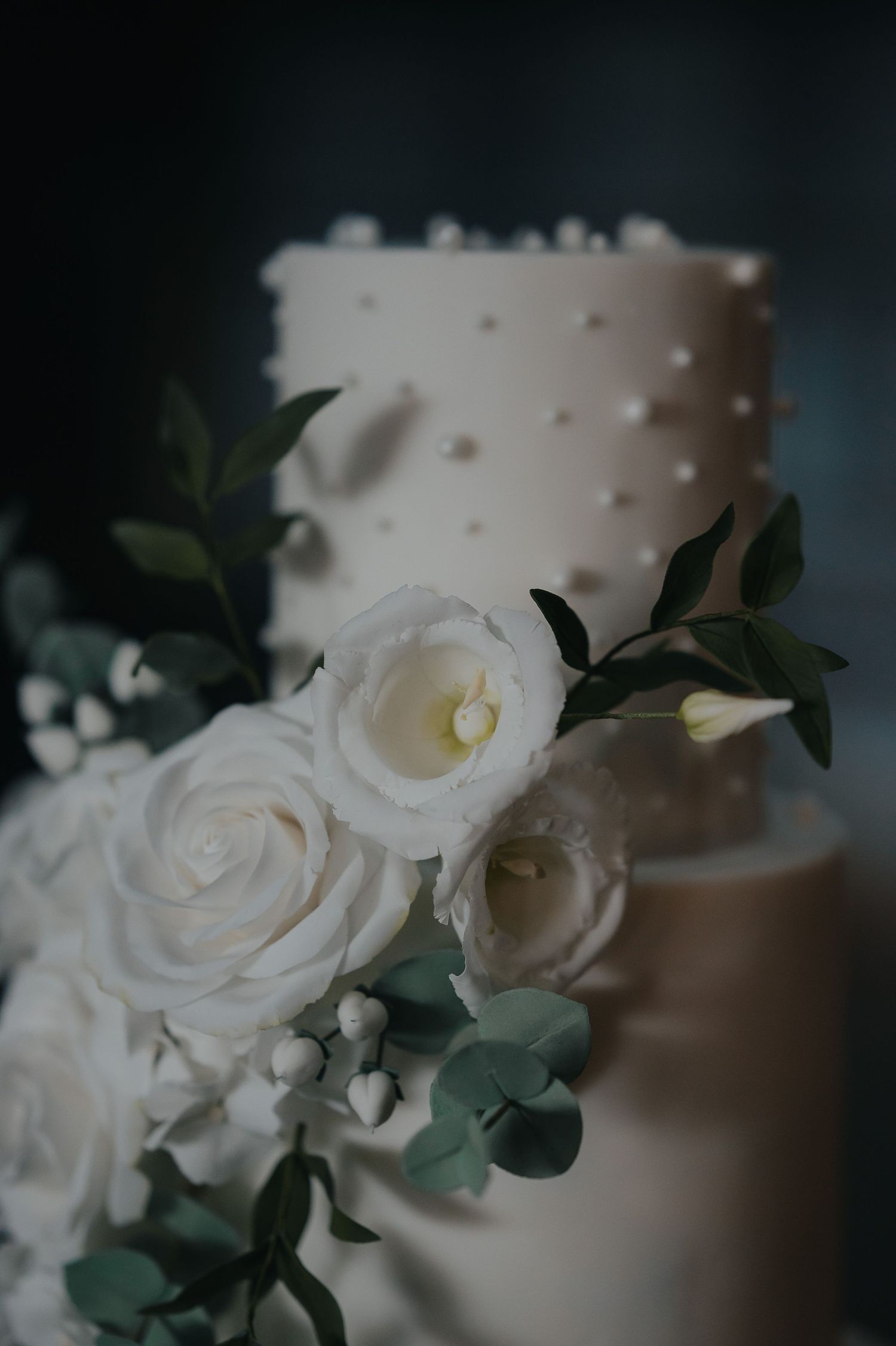 Elegant white wedding cake decorated with white roses and green foliage on a dark background.