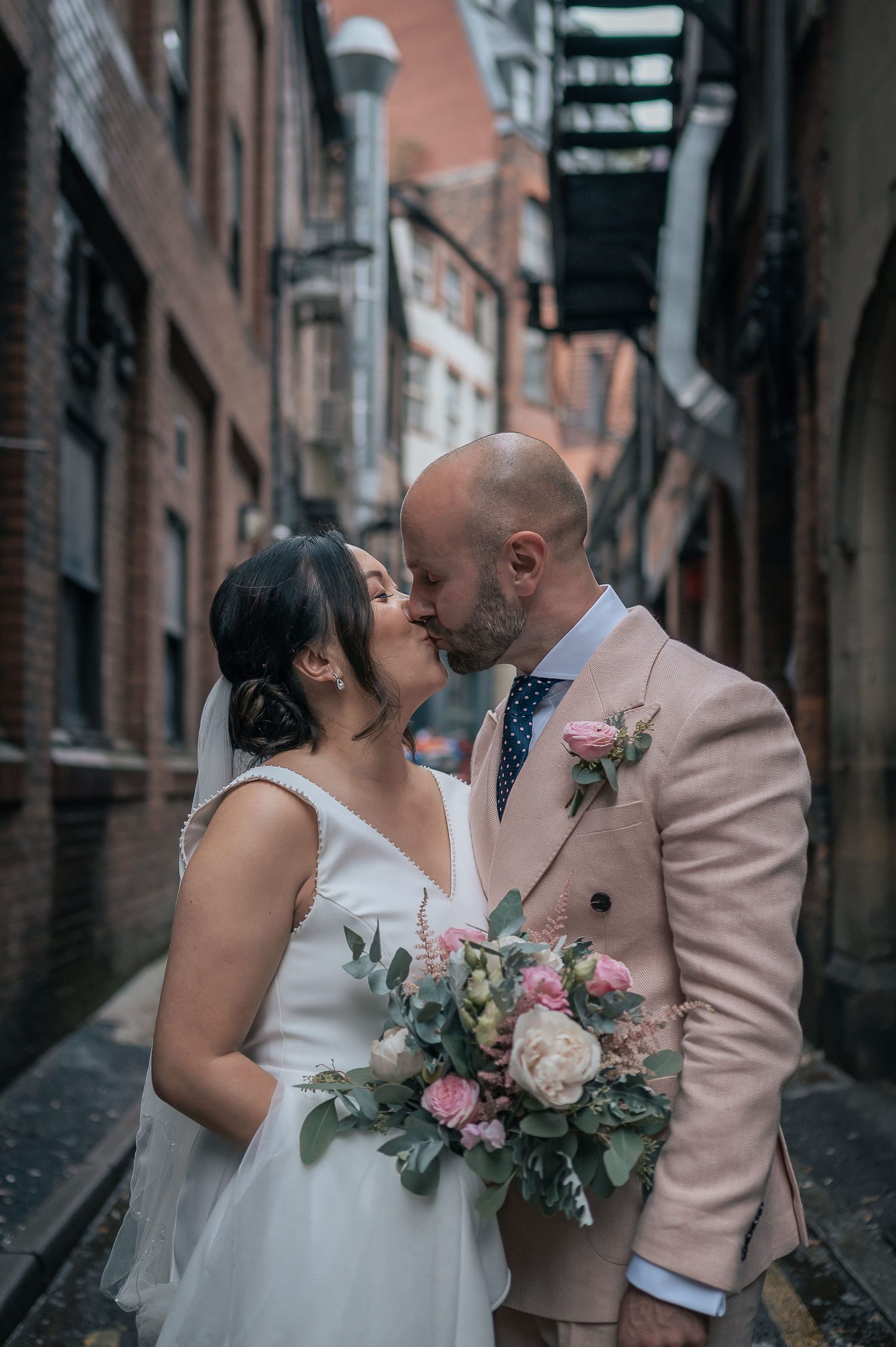 A wedding couple shares an intimate moment in a brick alleyway with a pink floral bouquet.