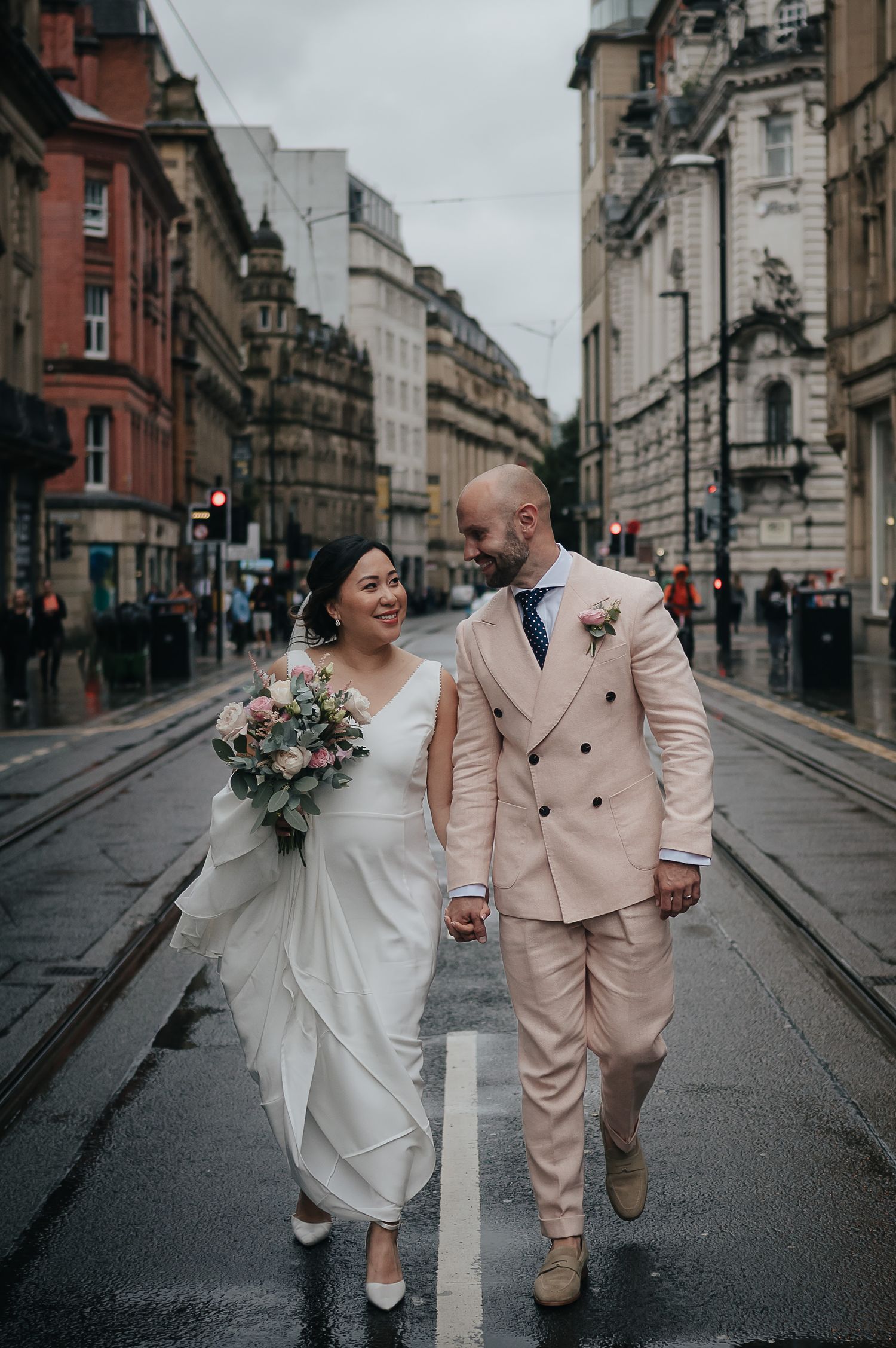 Couple walking down rainy city street in wedding attire against historic architecture.