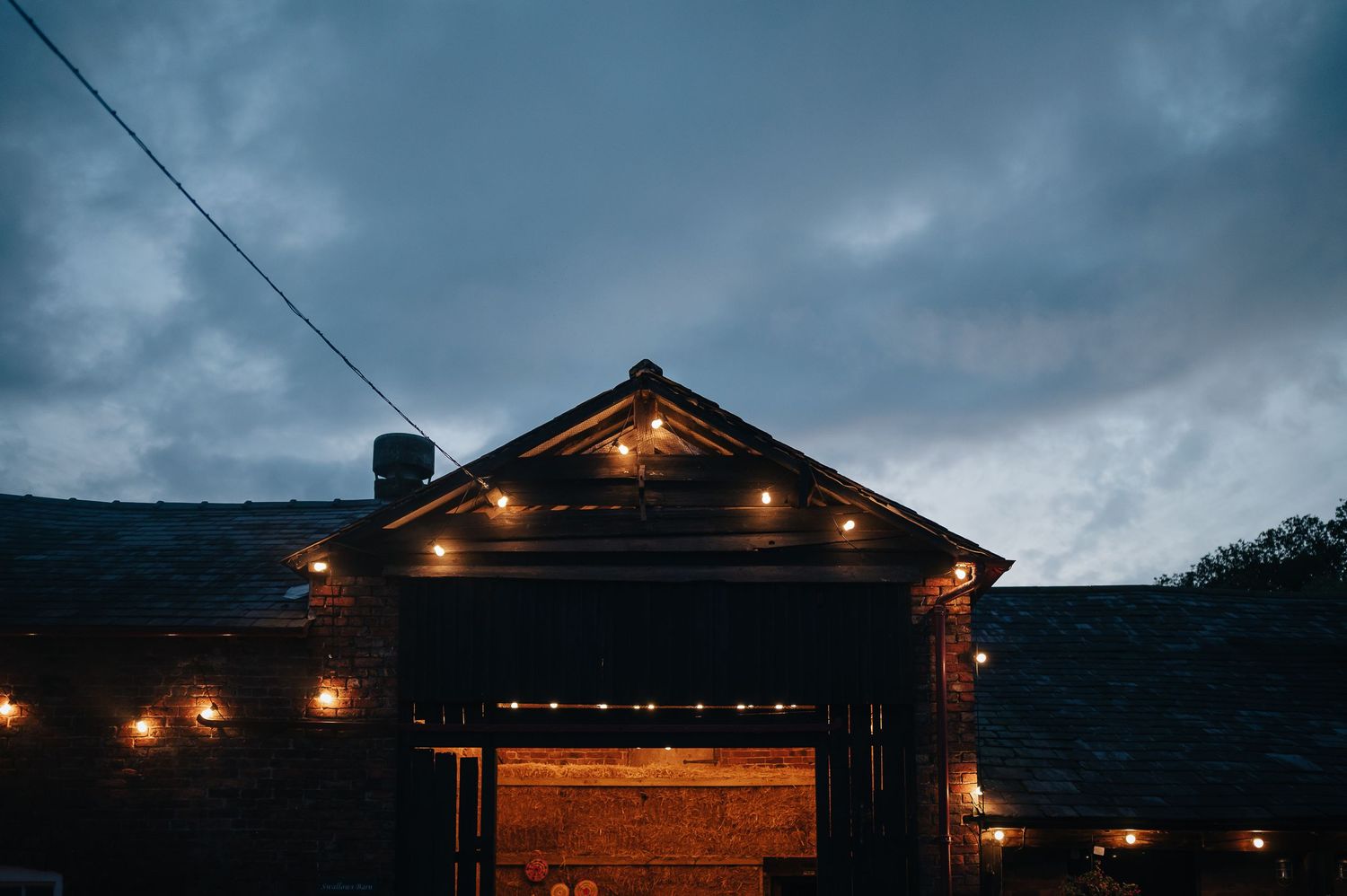 A rustic barn venue at dusk with string lights illuminating the roofline against cloudy skies.