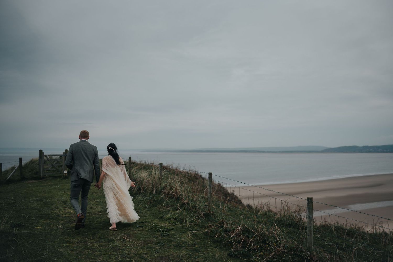 A couple walks along a coastal cliff path overlooking a vast beach under moody skies.