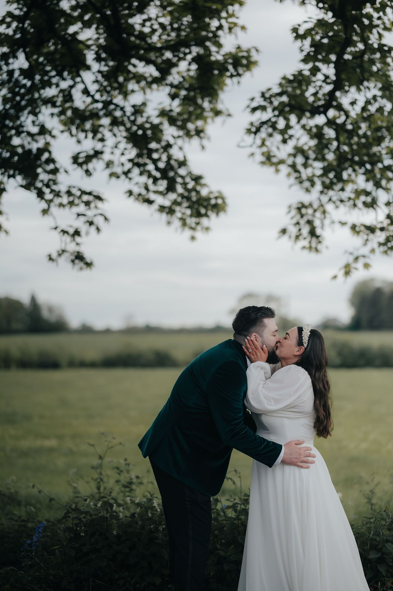 Romantic wedding moment outdoors with green trees and fields in background.