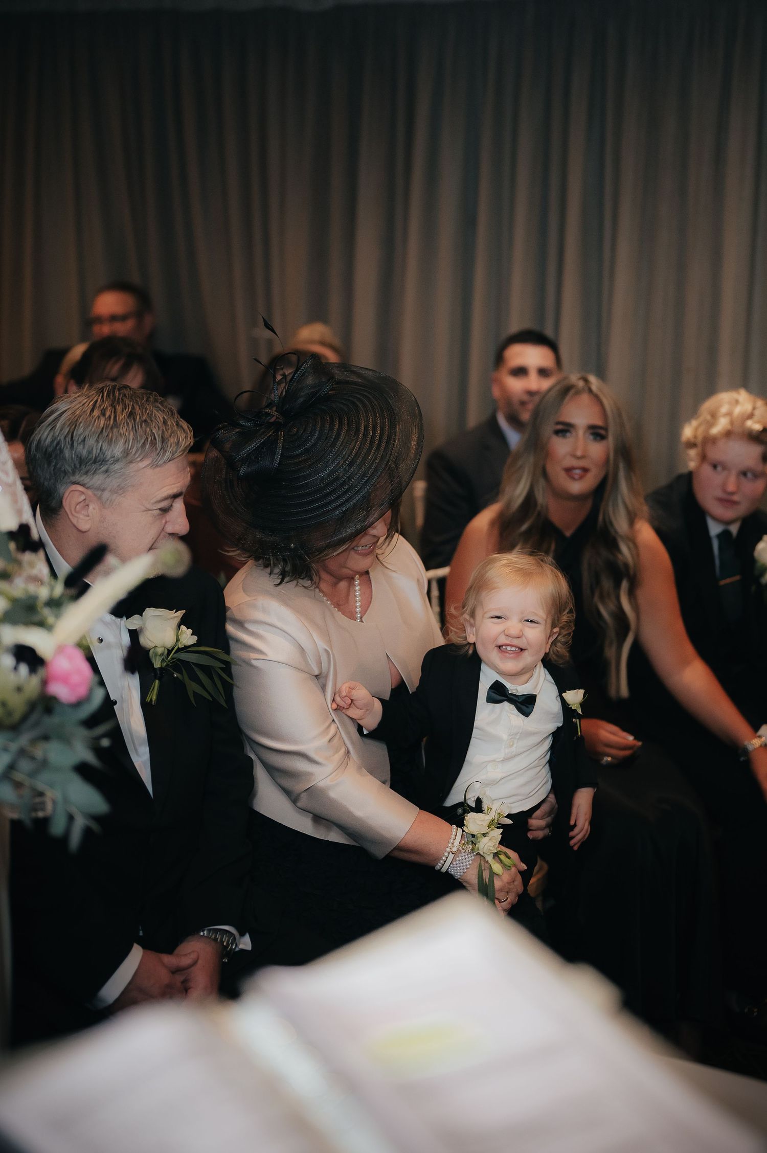 A smiling young child in a black tuxedo and bowtie sits among wedding guests during a ceremony.