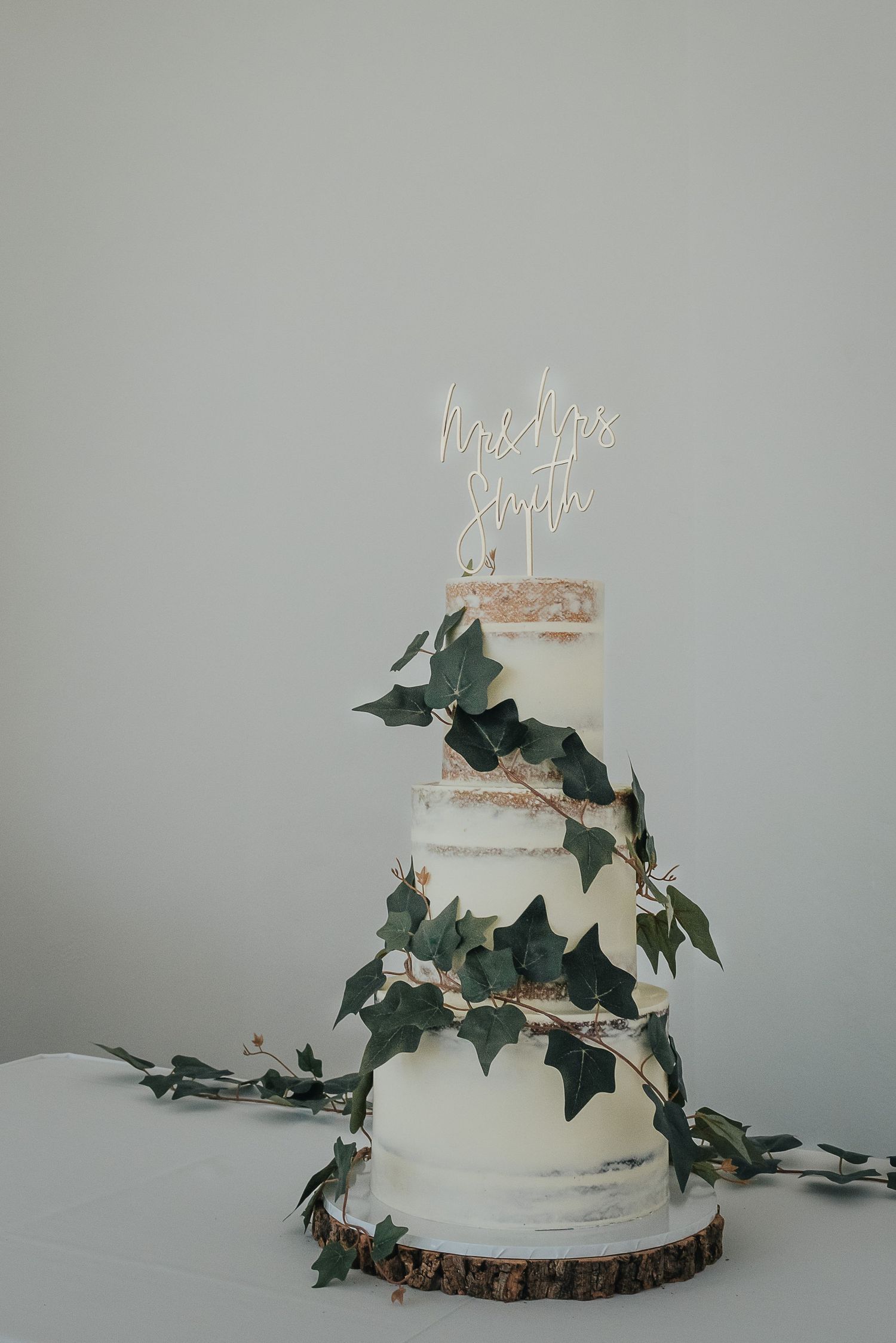 Three-tiered white wedding cake decorated with cascading ivy leaves and rustic wooden base.