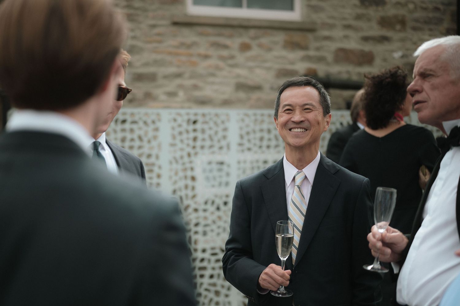 Wedding guests in formal attire gather outdoors for a social reception at a stone building venue.