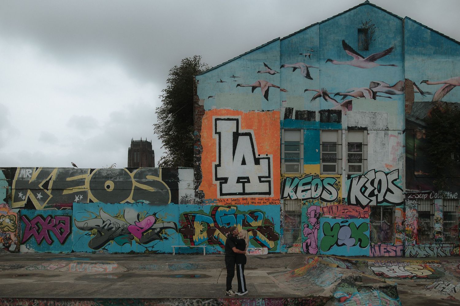 Graffiti adorns a turquoise building wall with birds painted above and a lone figure standing in front of urban art.