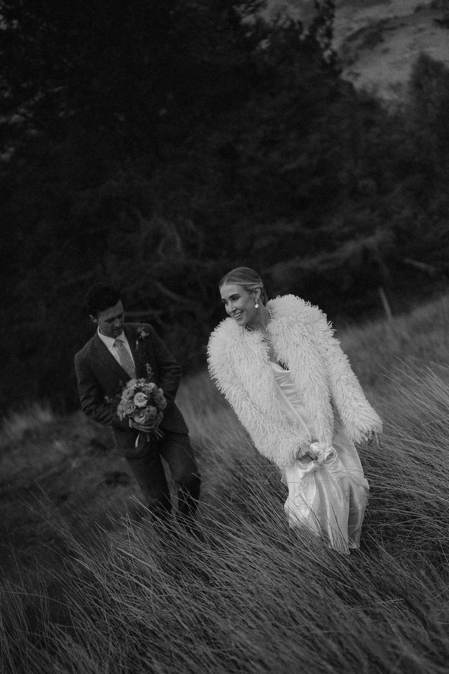 Black and white photo of a bride in fur coat and groom holding bouquet walking through tall grass outdoors.
