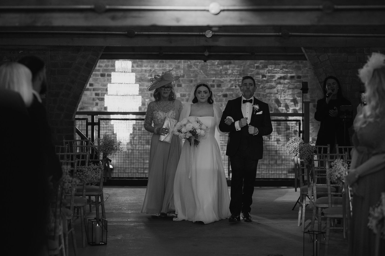 A wedding ceremony procession in a rustic brick venue with guests seated along the aisle.