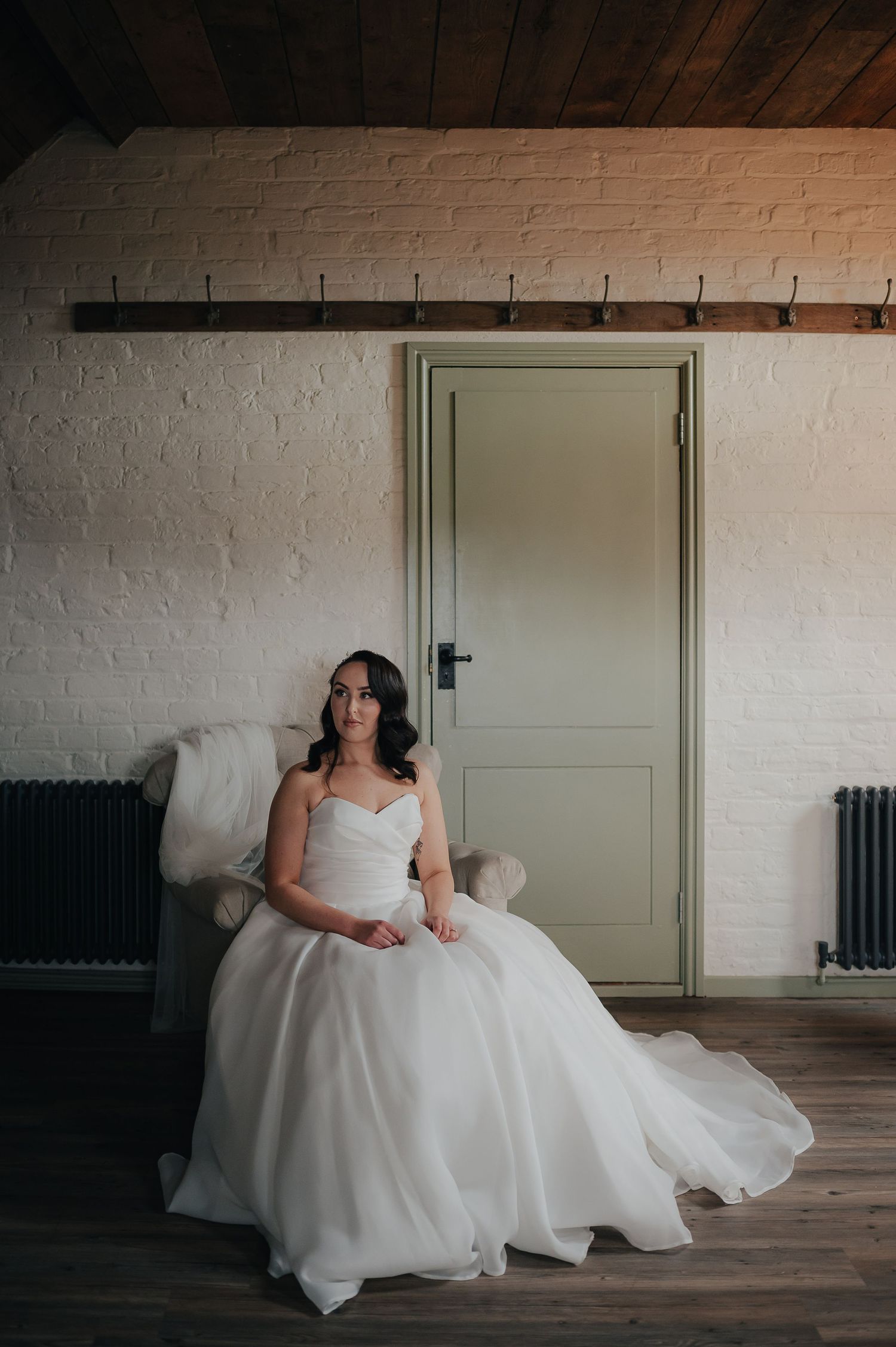Bride in flowing white gown sitting elegantly against white brick wall with green door.