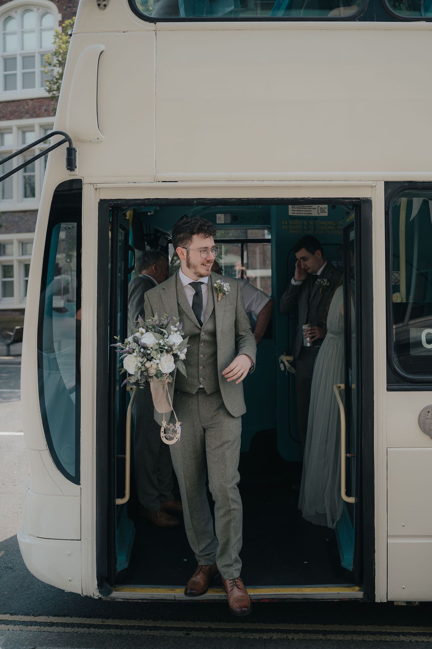 A person in a grey suit stepping off a white double decker bus while holding a floral bouquet.