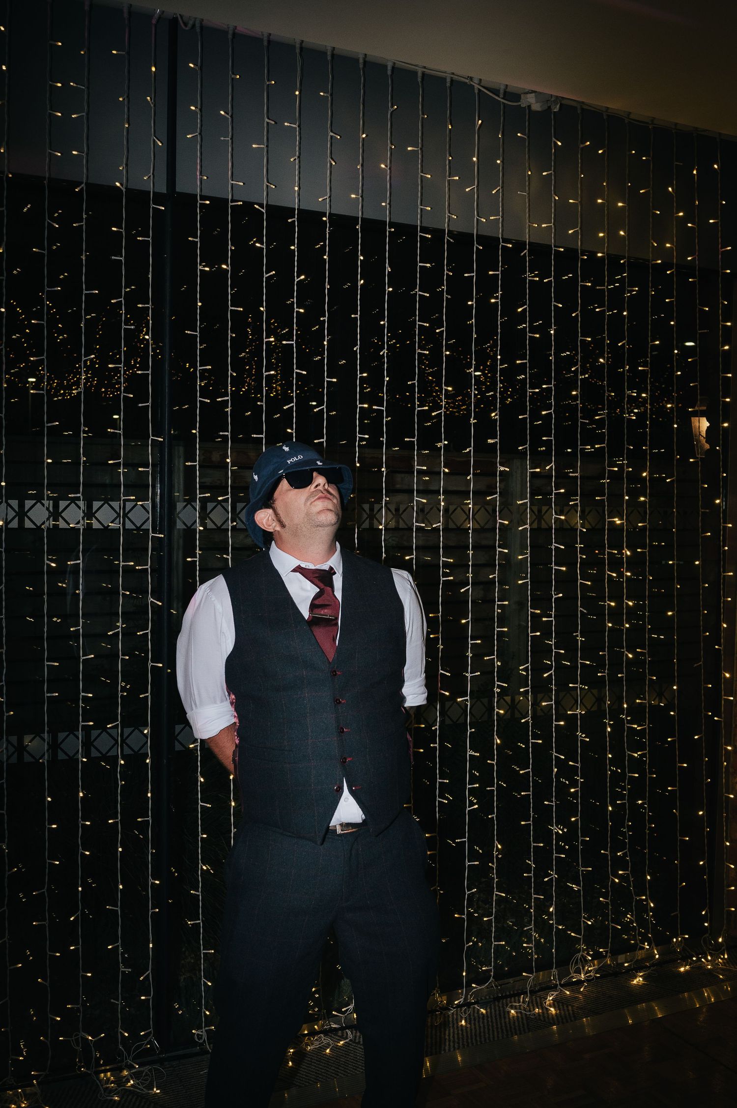 Guest in formal attire posing against wall of cascading fairy lights at wedding reception.