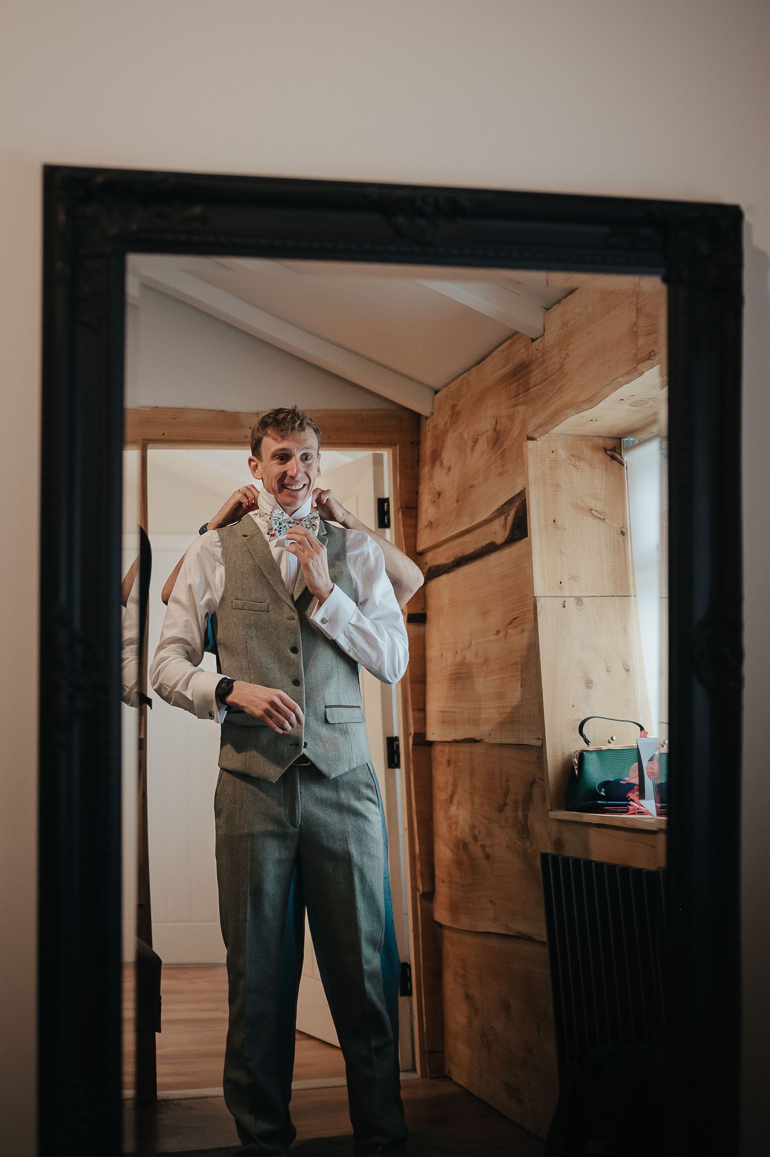 A groom in a gray three-piece suit prepares for his wedding in a rustic wooden doorway.