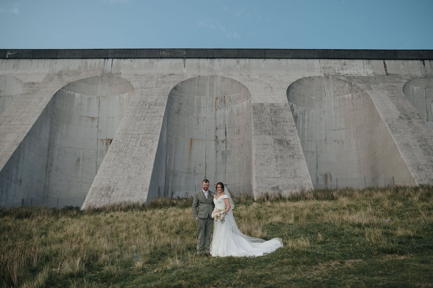 Newlyweds pose against massive concrete dam structure under blue sky.