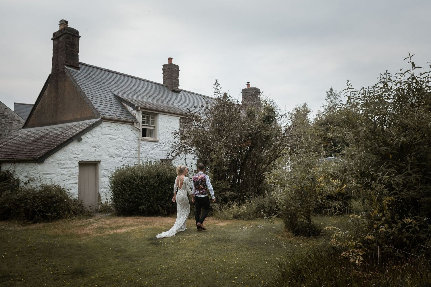 A couple walks hand in hand near a white cottage house on an overcast day.
