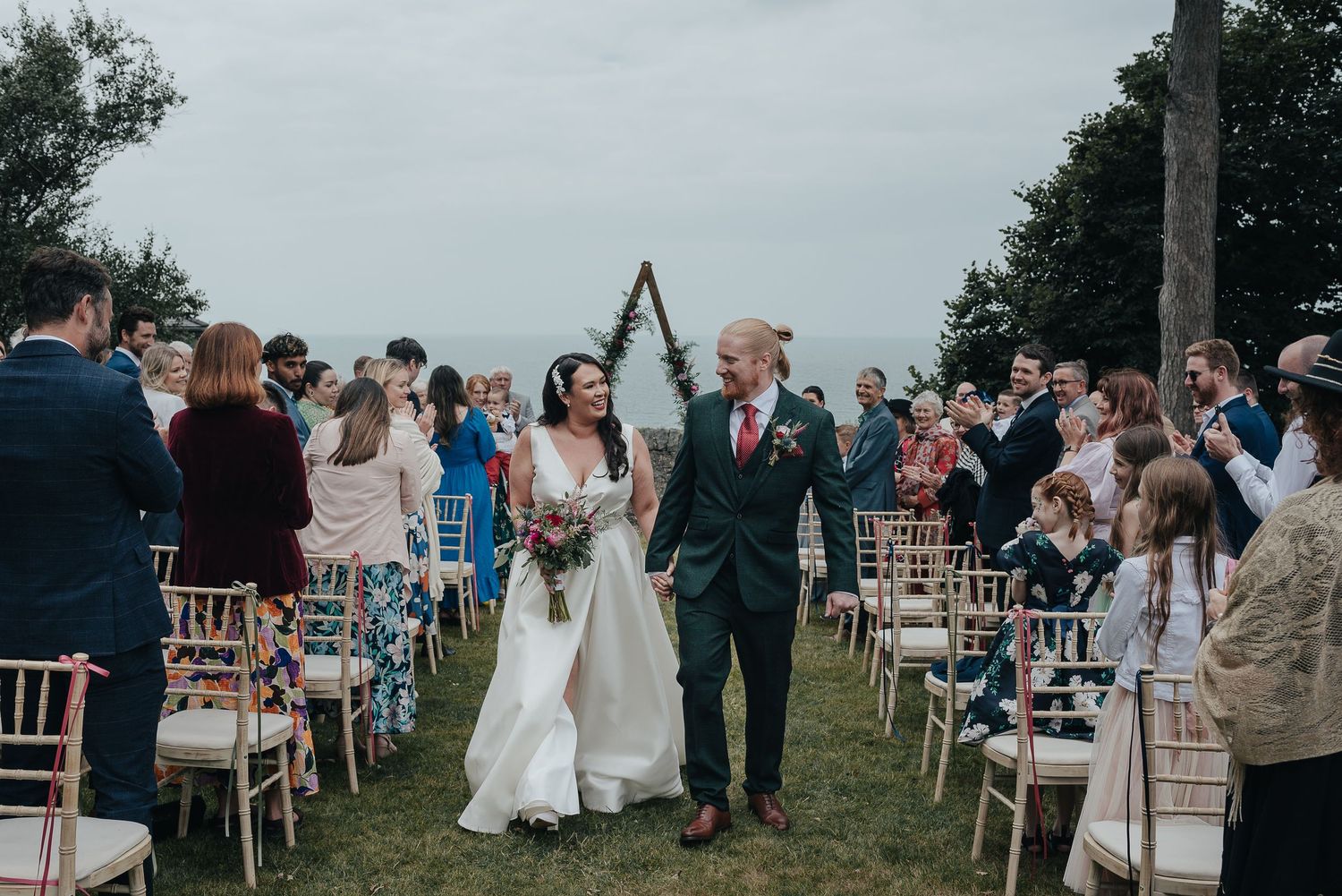 Wedding couple walking down outdoor aisle between rows of wooden chairs.