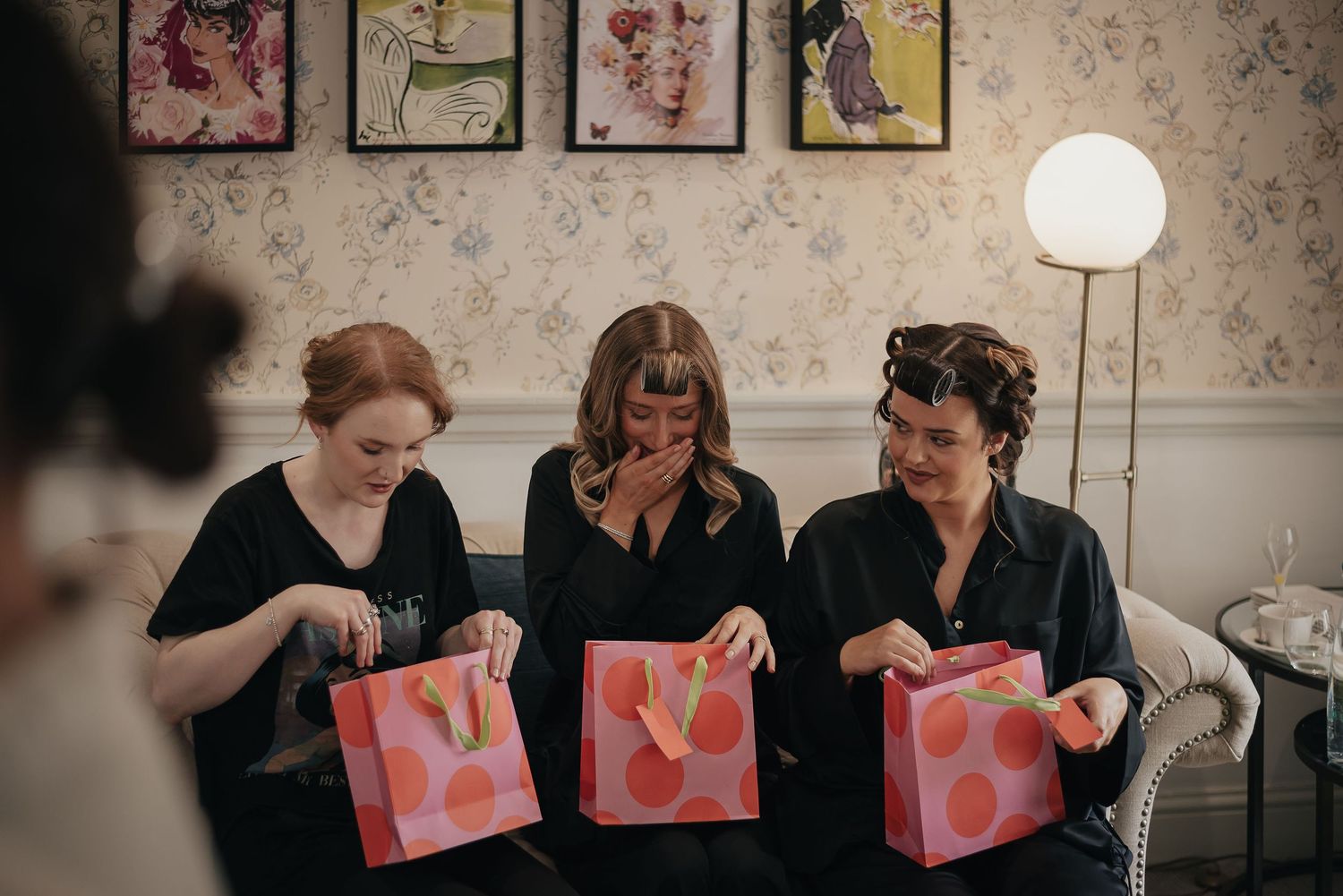 Three guests sitting on couch opening pink gift bags in decorated room.