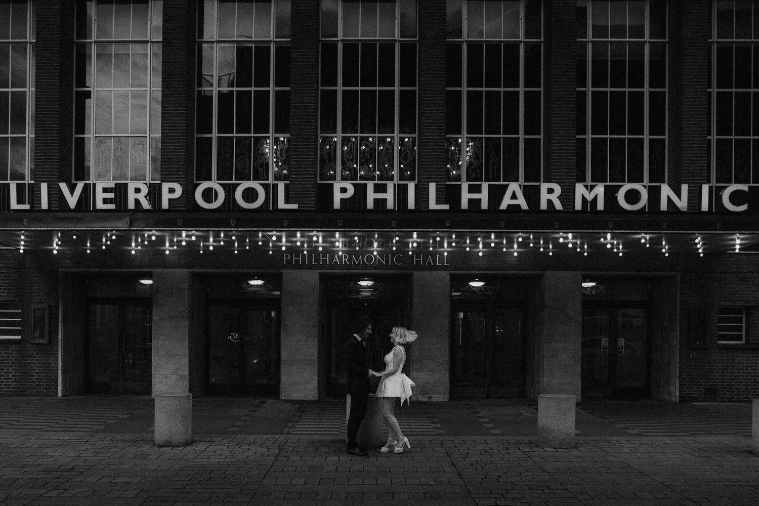 Black and white photo of a couple dancing outside the Liverpool Philharmonic Hall at night.