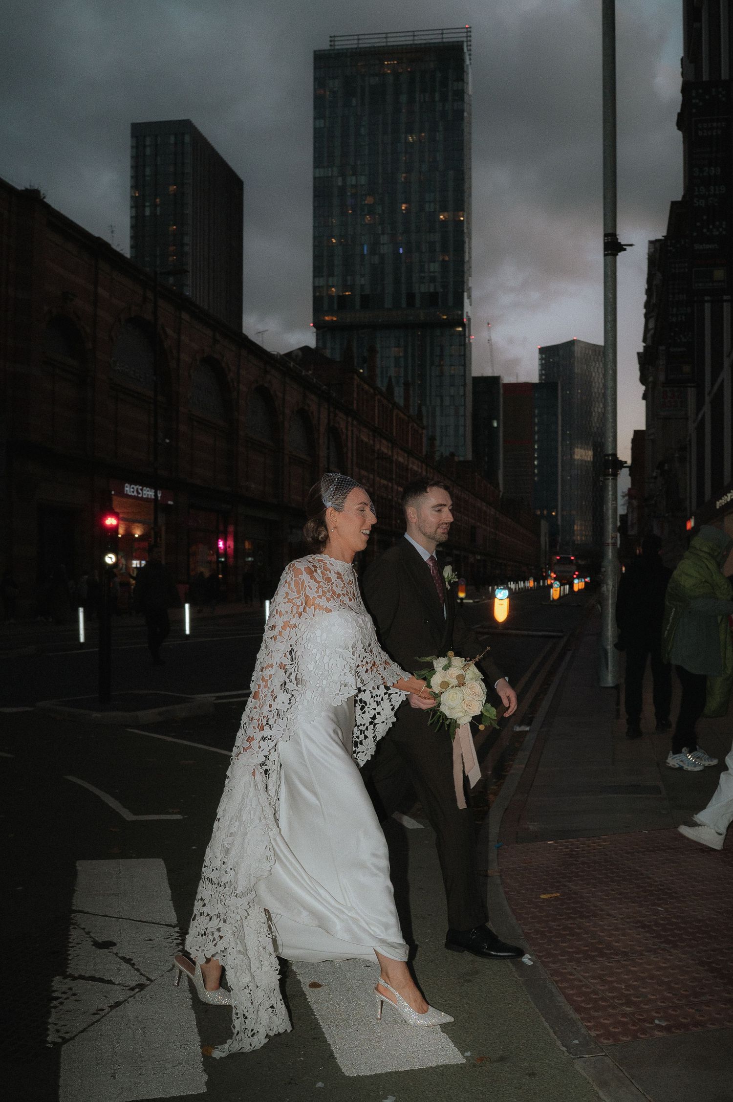 A couple in formal wedding attire walking along a city street at dusk with tall buildings in the background.
