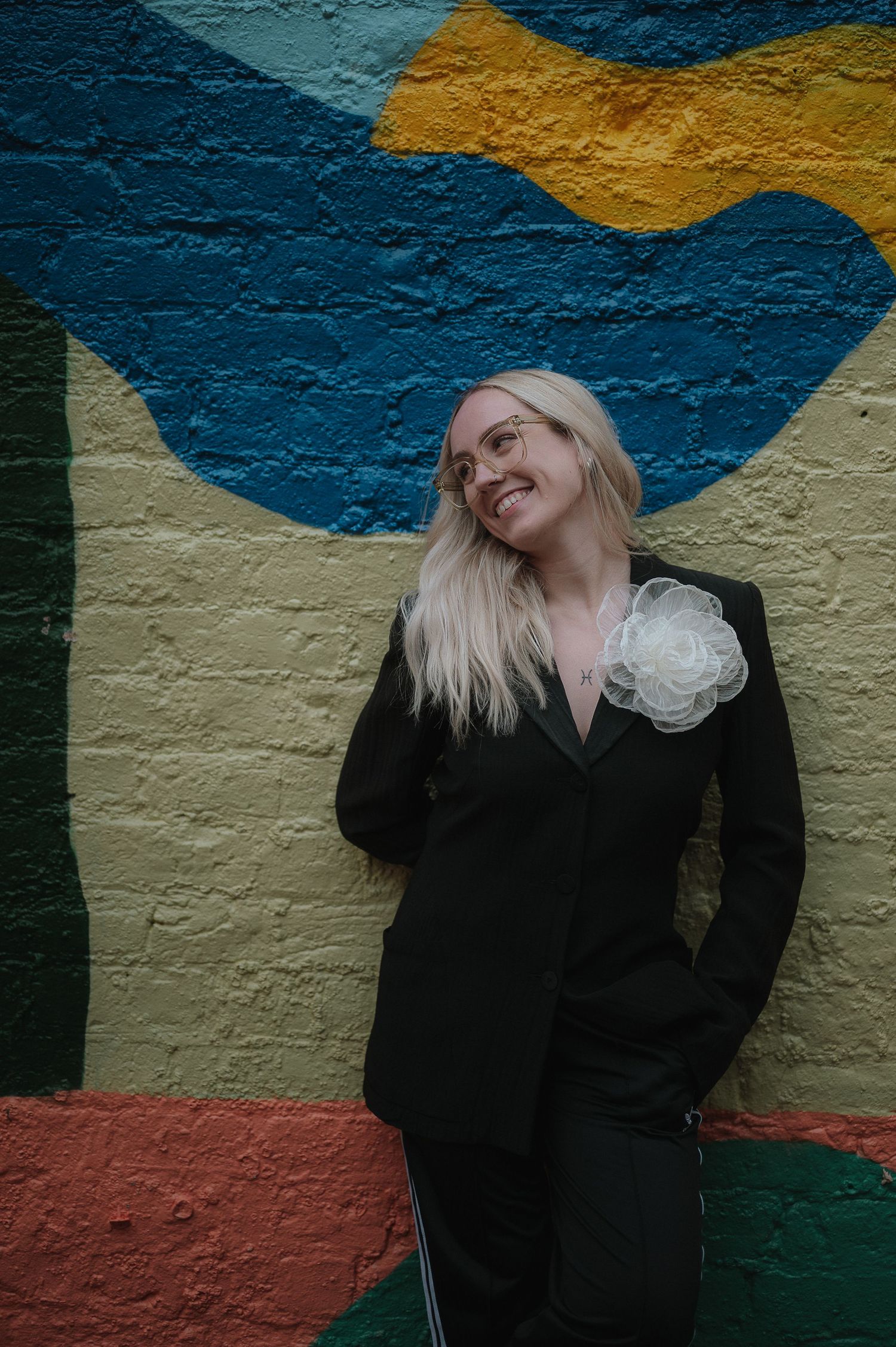 A person in black clothing poses against a colorful mural wall featuring blue and yellow abstract shapes.