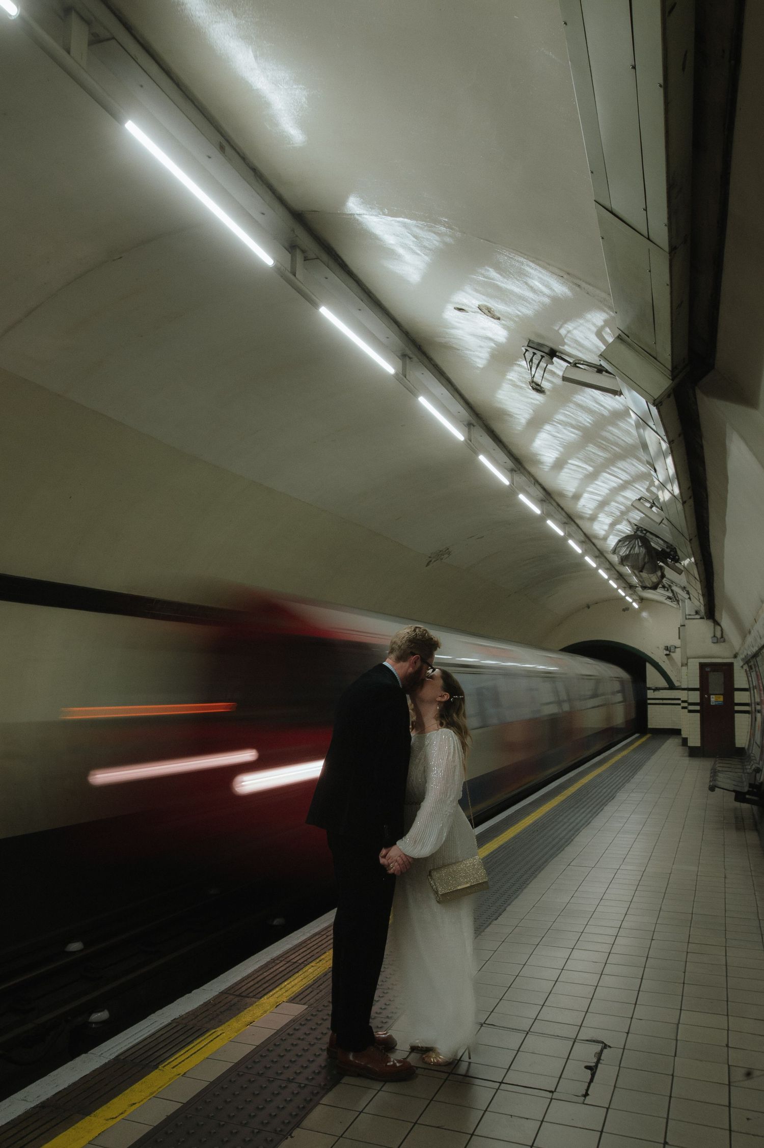 A couple waits on a subway platform in wedding attire with motion-blurred train passing.