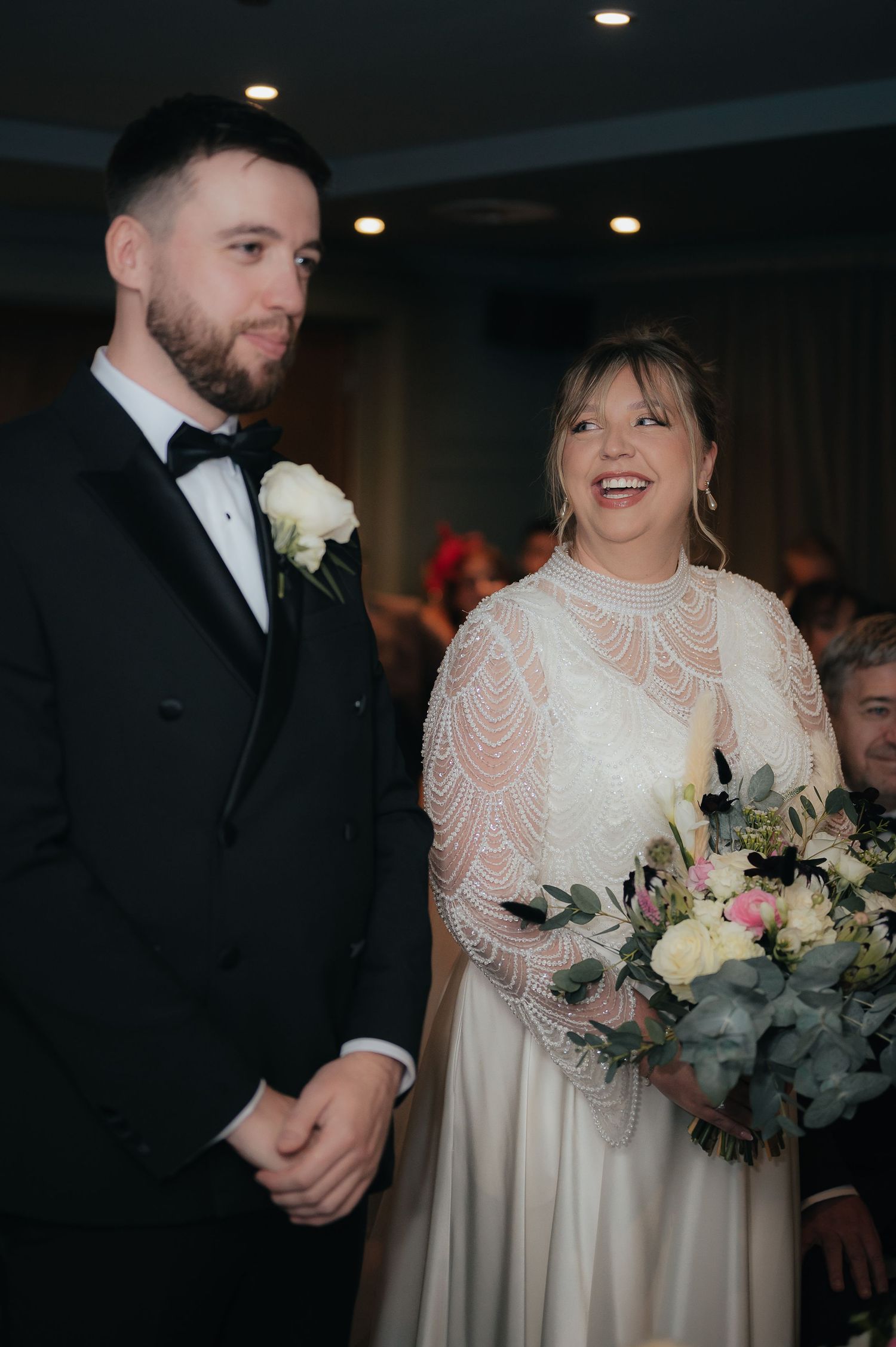 A couple shares a joyful moment during their wedding celebration while holding a floral bouquet.