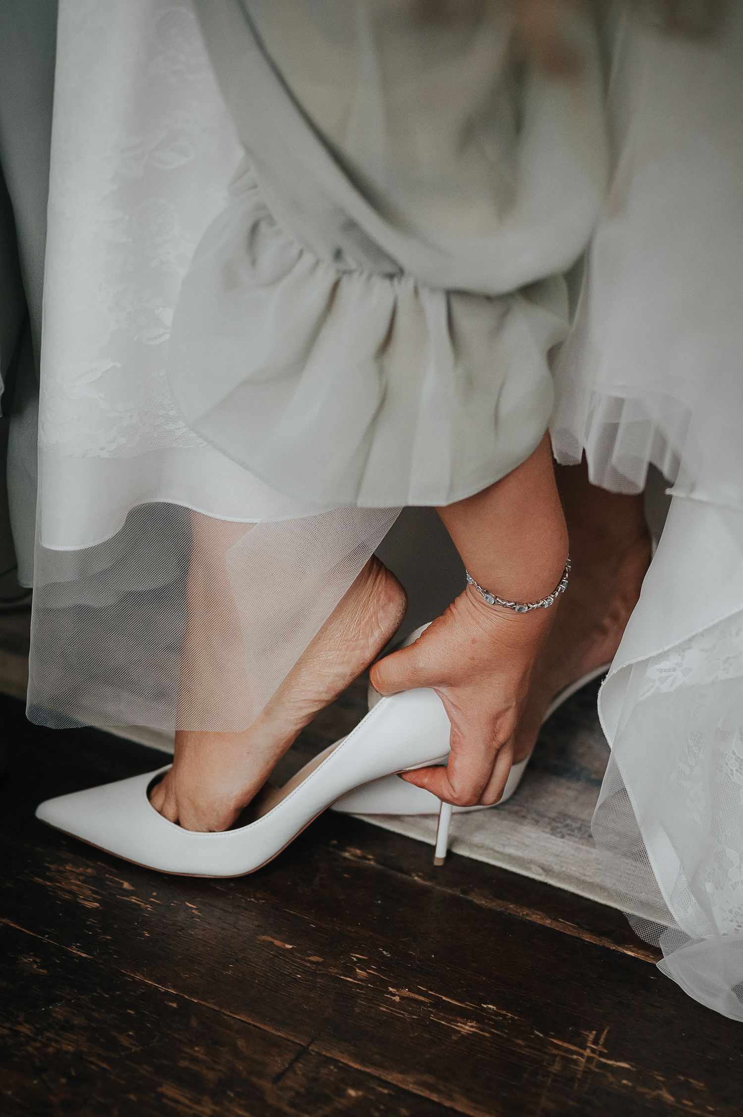 Close-up of white stiletto heel and anklet being put on against wooden floor.