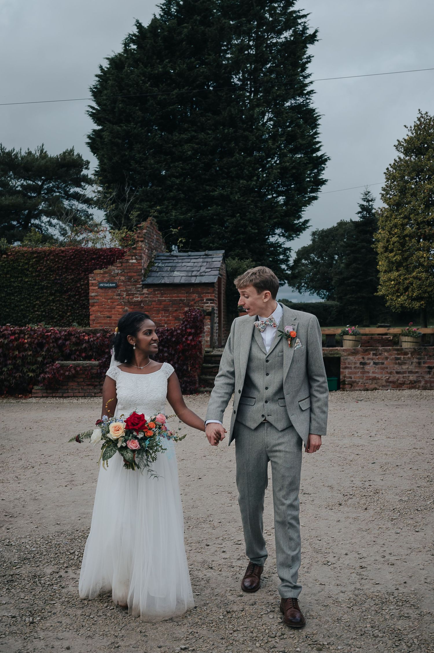 A couple walking hand in hand on a gravel path by a brick building with autumn foliage.
