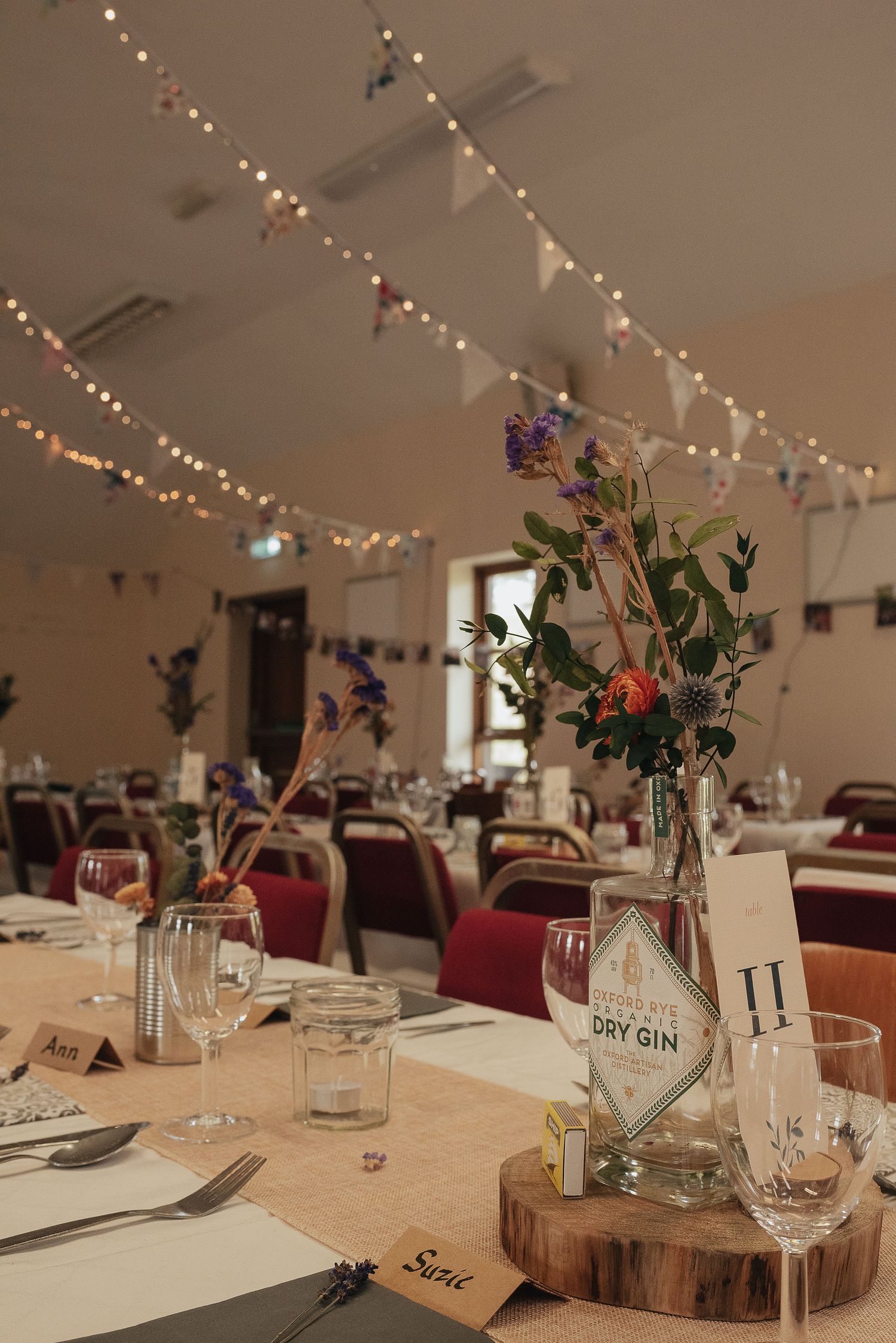 Wedding reception table setting with fairy lights and floral centerpiece featuring gin bottle vase on wooden slice.