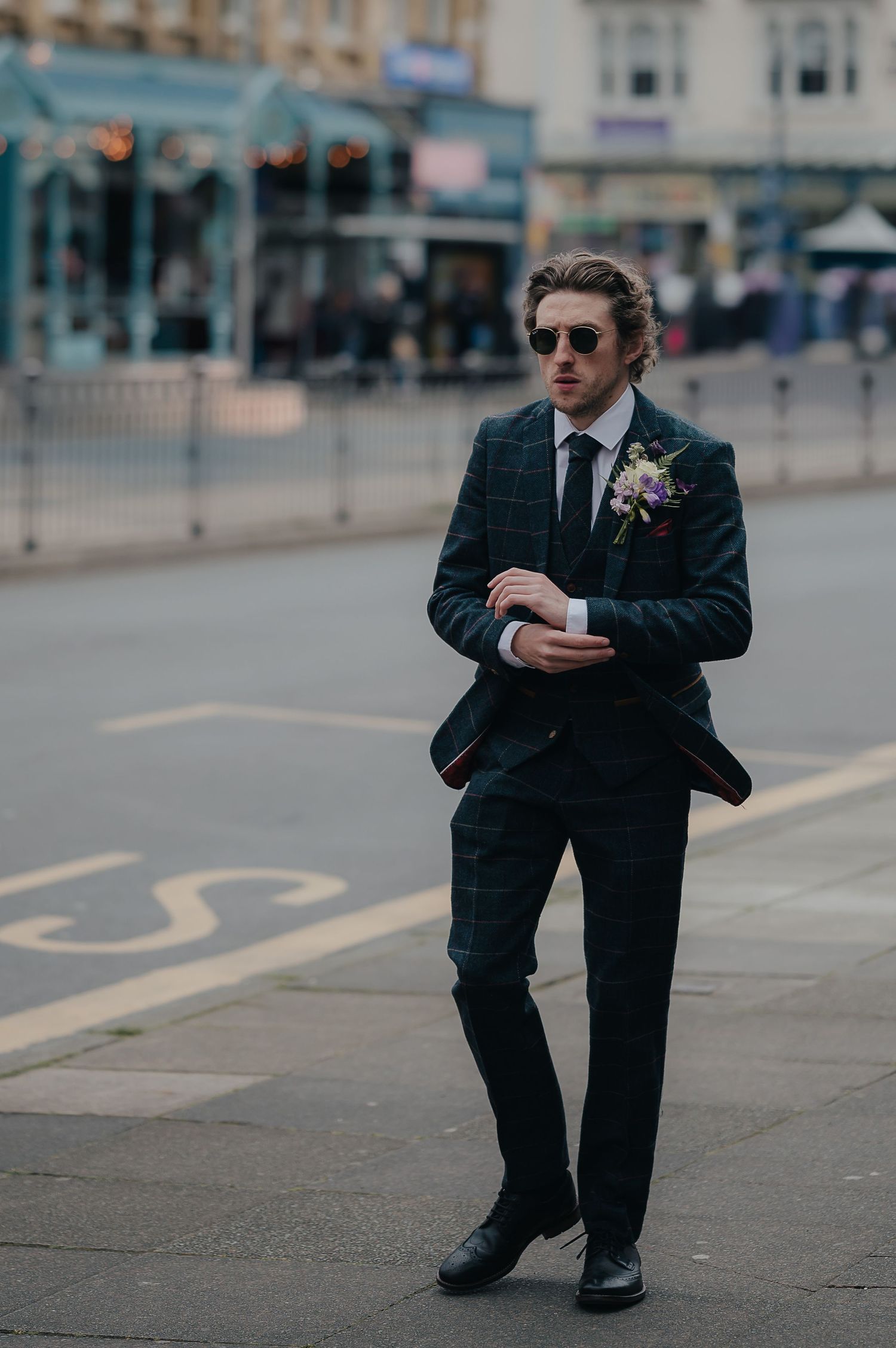 A person in a dark suit and sunglasses standing on a city sidewalk with a boutonniere.