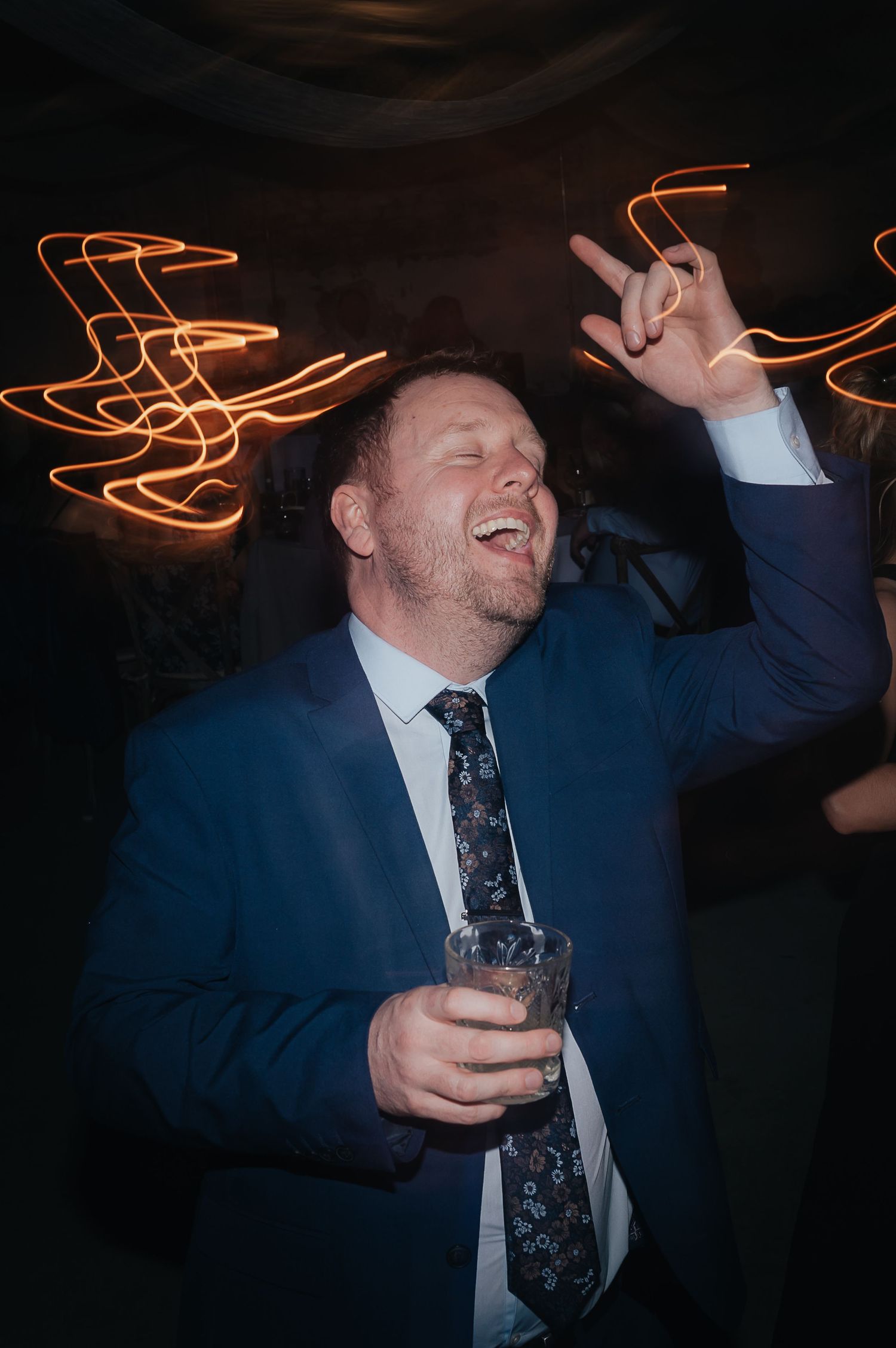 Guest in blue suit jacket celebrating at party with light trails and drink in hand.