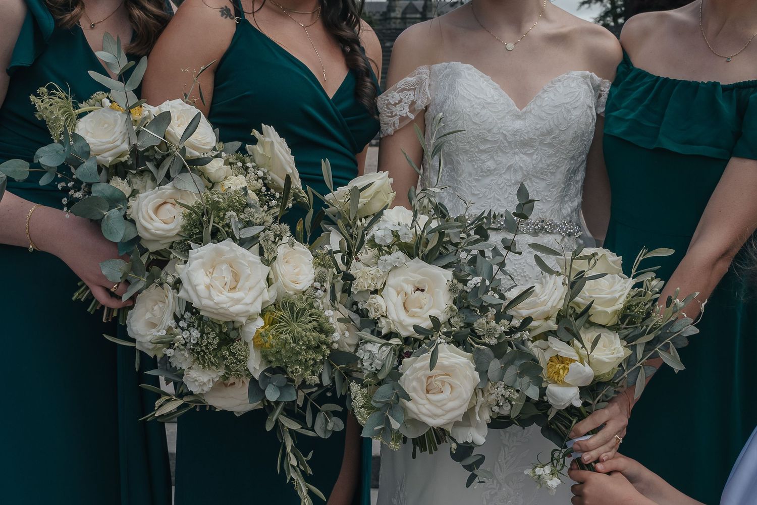 Elegant white rose bouquets with greenery held by wedding party wearing emerald green dresses.