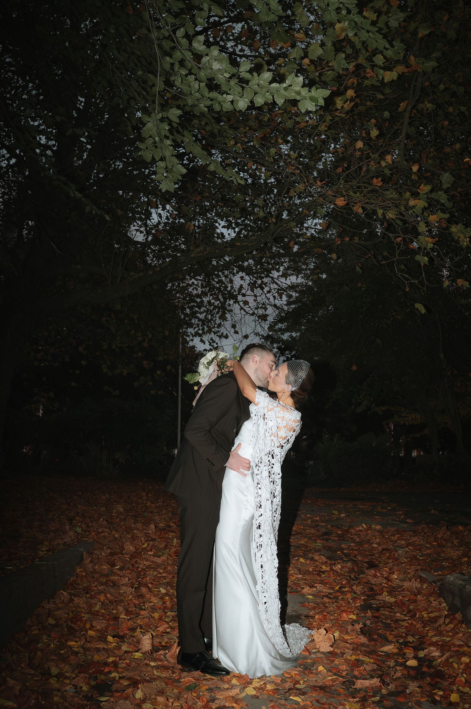Bride and groom share a romantic kiss outdoors at night, surrounded by autumn leaves and trees in a wedding photo.