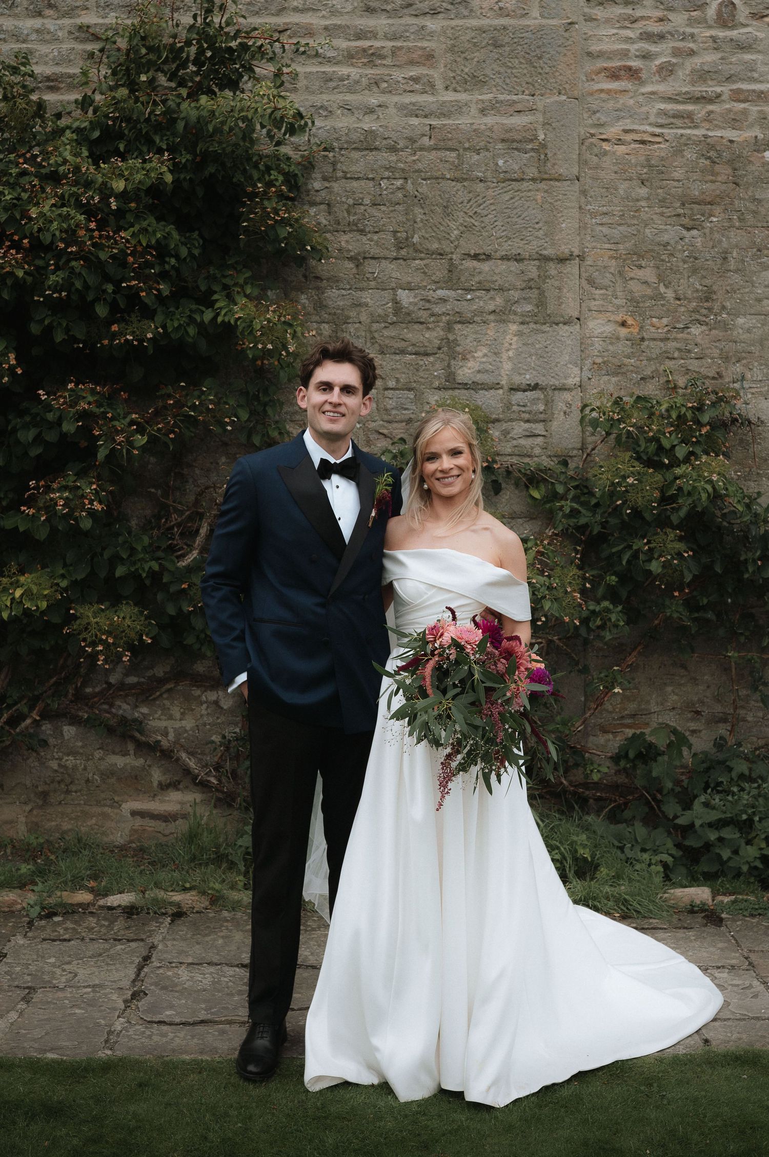 A wedding portrait against a stone wall with lush greenery.