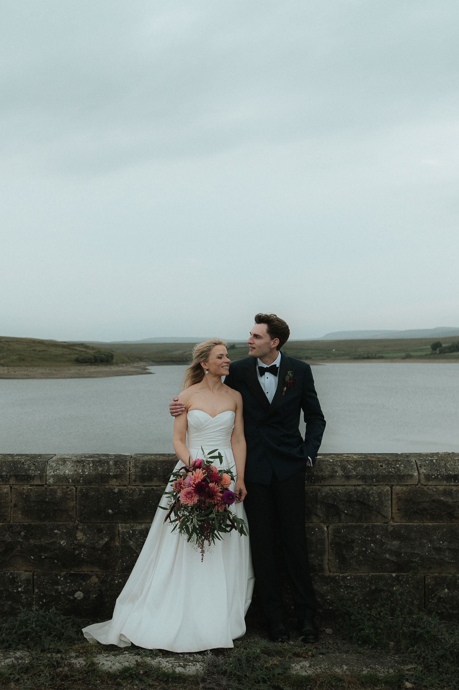 A couple in wedding attire share intimate moments by a stone wall overlooking a misty lake on their special day.